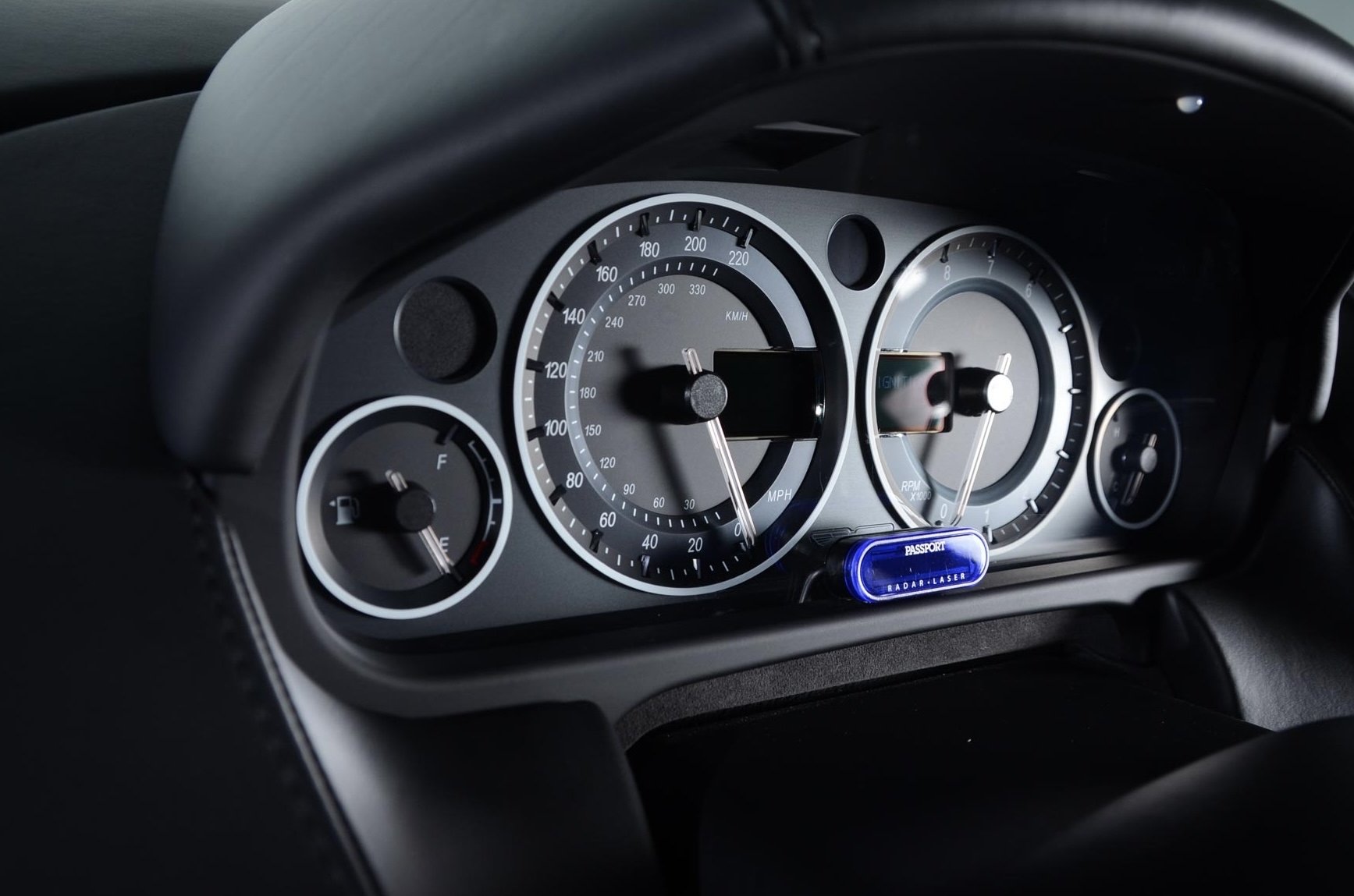 Close-up of a car dashboard showing speedometer, tachometer, fuel gauge, and a radar laser device with a blue light labeled 'PASSPORT'.