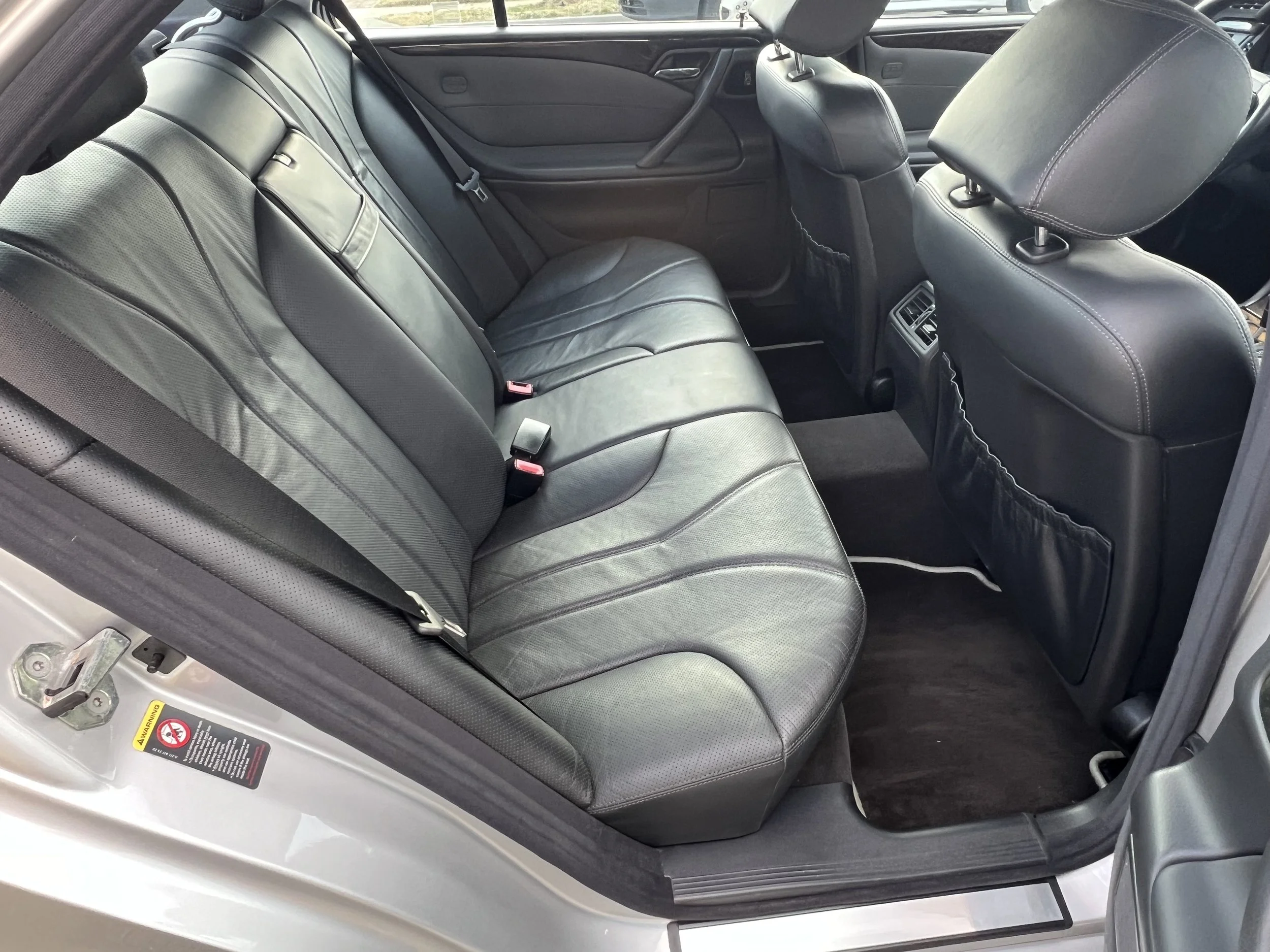 The interior of a car showing the backseat area with black leather seats, seat belts, and headrests. The front seats and part of the dashboard are also visible.