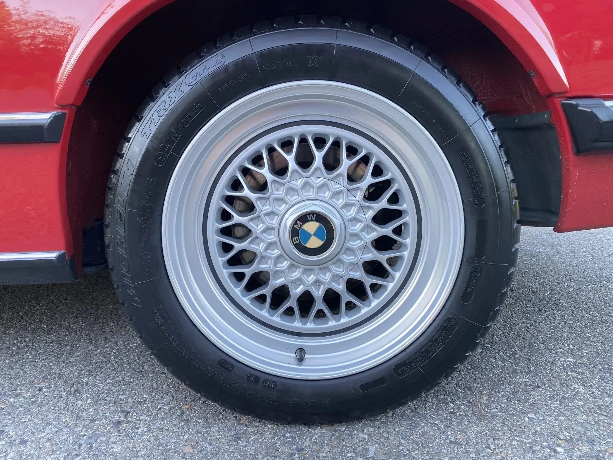Close-up of a BMW car wheel with a BMW logo in the center, featuring a multi-spoke alloy rim and a black tire, part of a red car body.