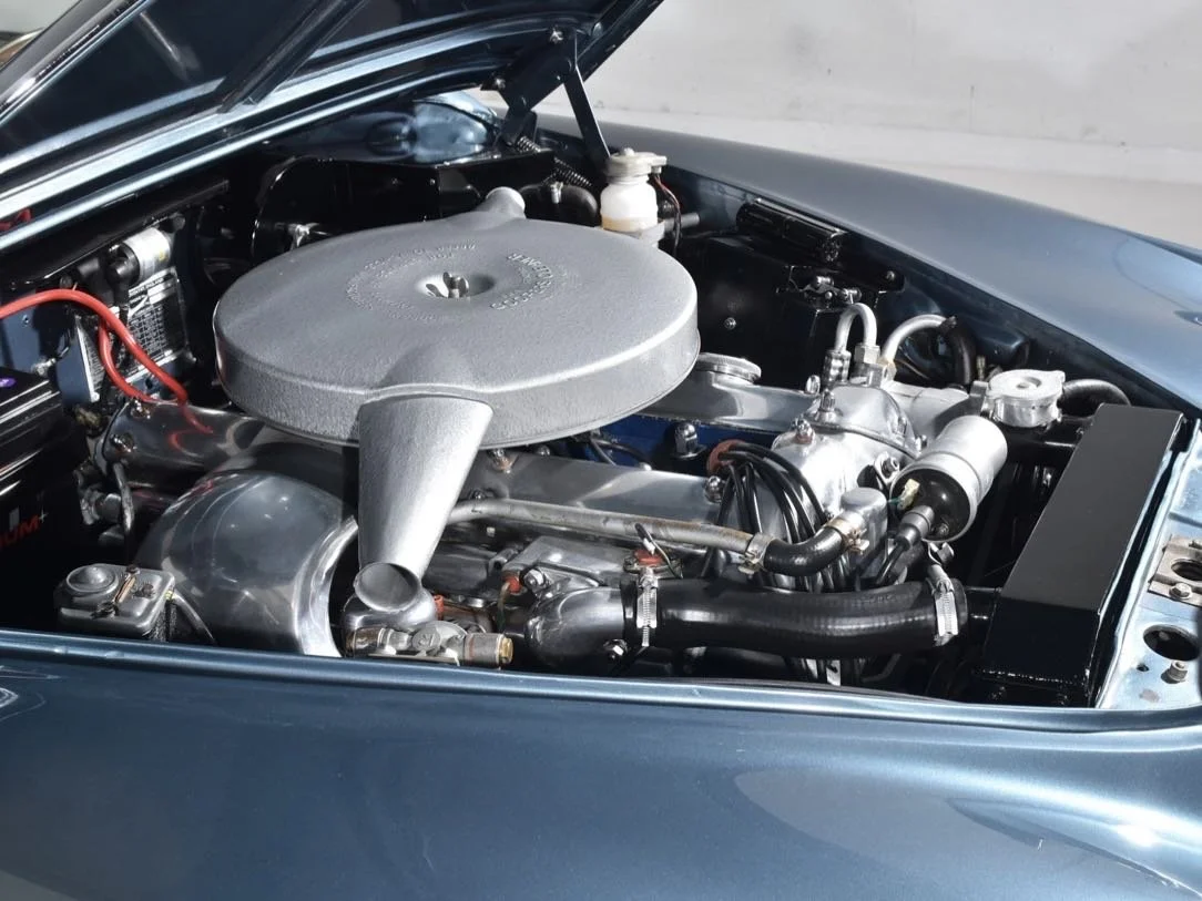 Close-up of a vintage car engine with metallic components, black hoses, and a large circular air filter on top.