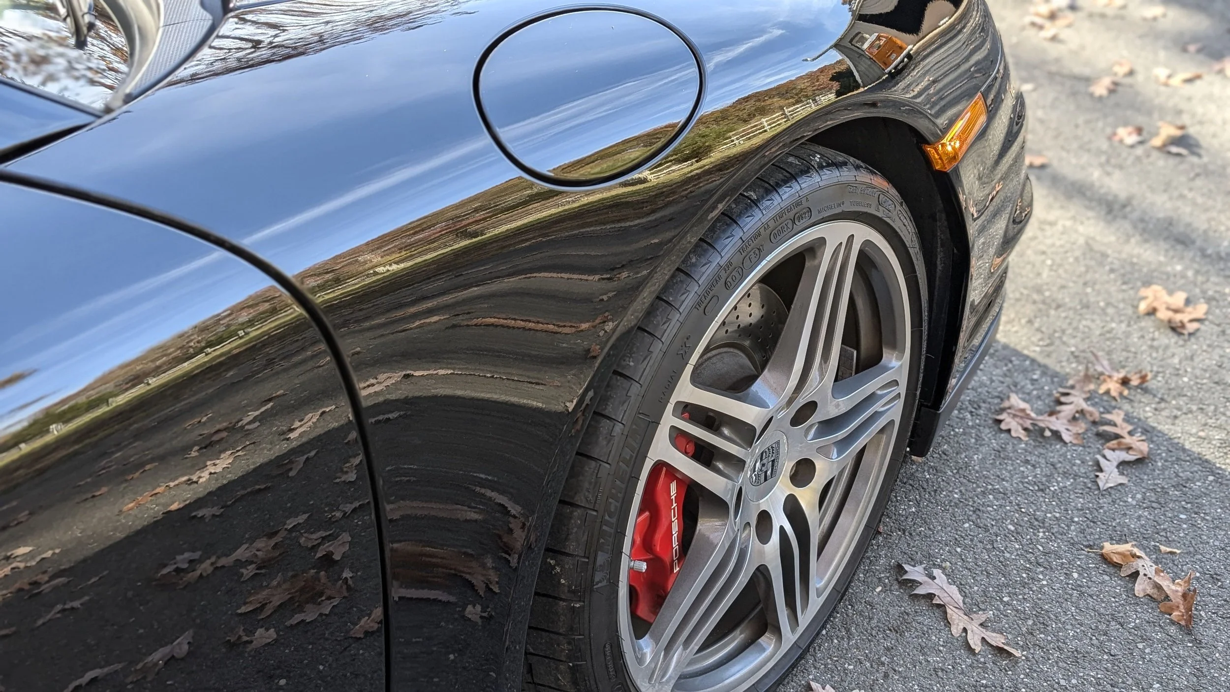 Close-up of a black sports car's front wheel, featuring a silver multi-spoke rim, red brake caliper labeled Porsche, and a low-profile tire, parked on a driveway with fallen leaves.