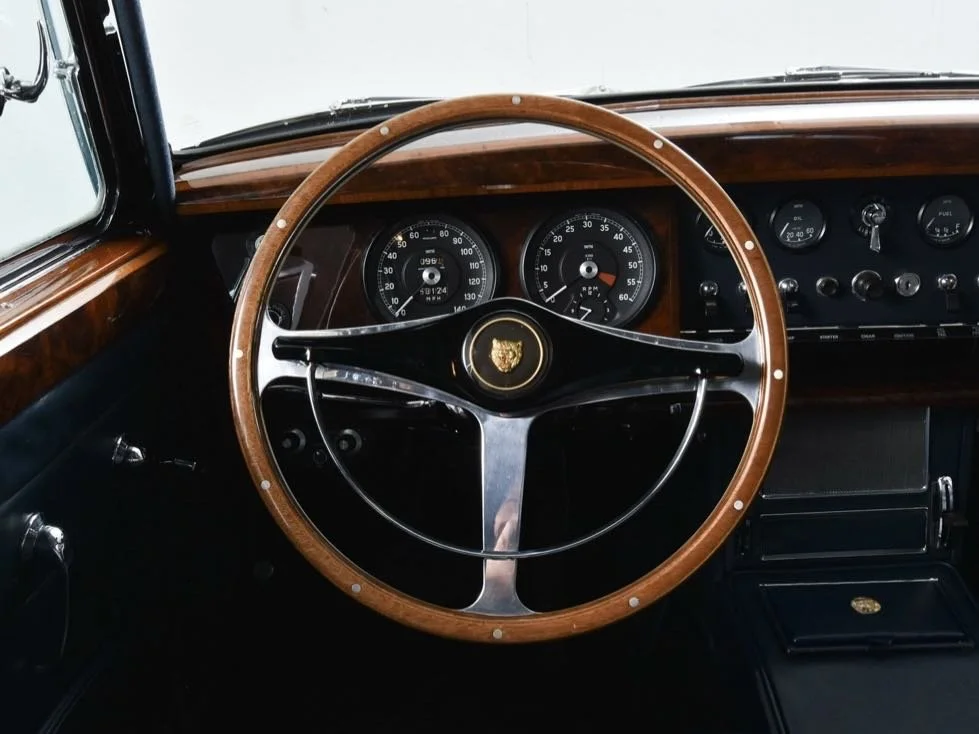 Interior of a classic car showing a wooden steering wheel with a gold crest in the center and a dashboard with analog gauges.