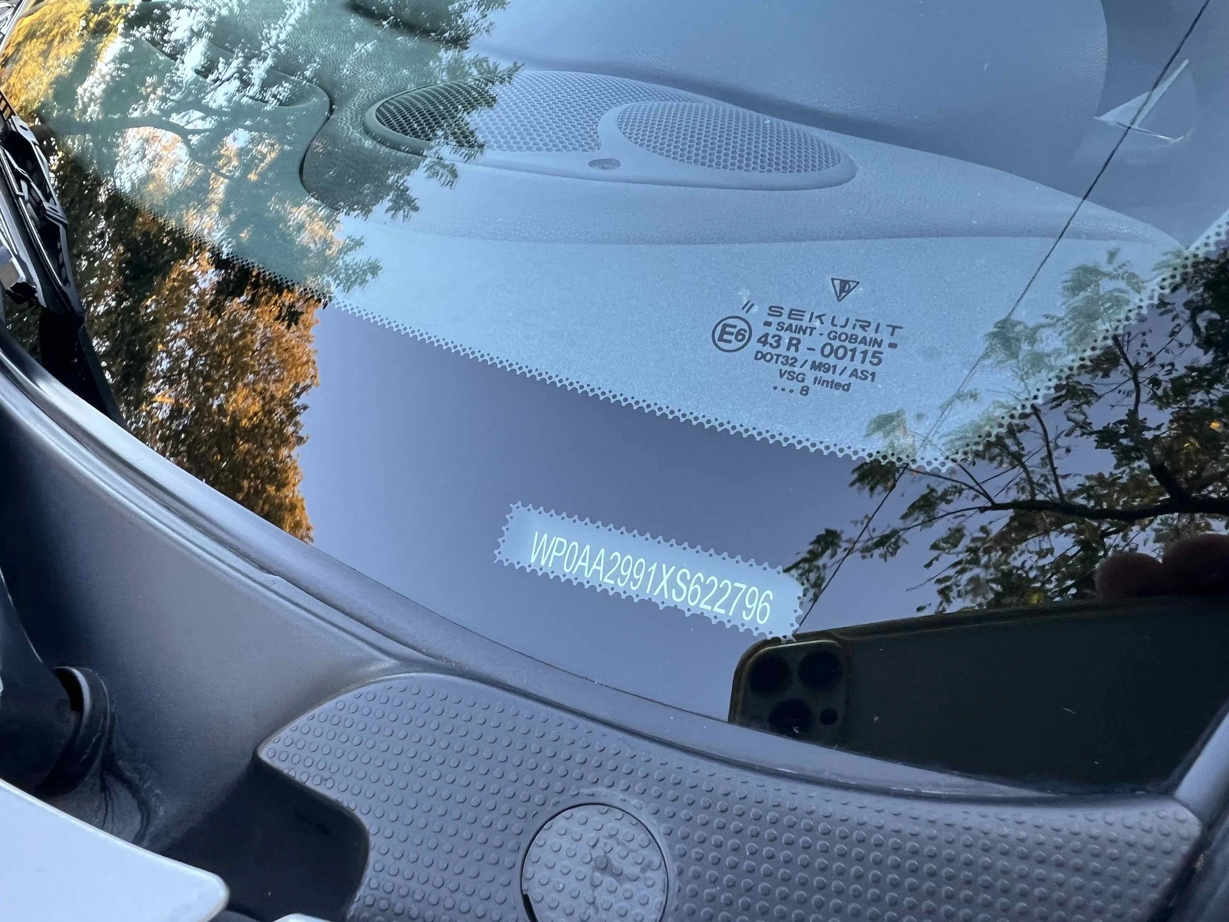 A close-up photo of a car's windshield displaying a Vehicle Identification Number (VIN) with reflections of trees and sky, and a portion of the dashboard visible.