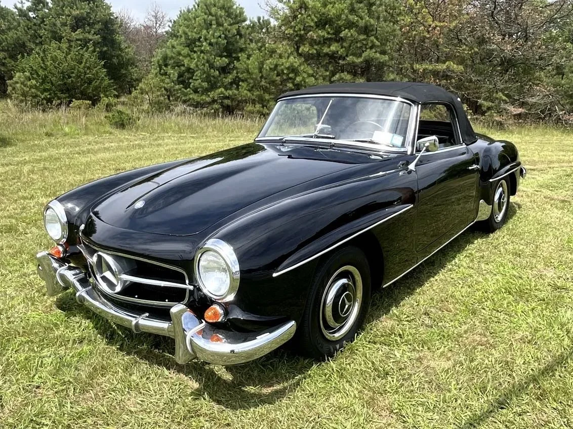 A vintage black Mercedes-Benz convertible with a black soft top parked on grass, surrounded by trees.