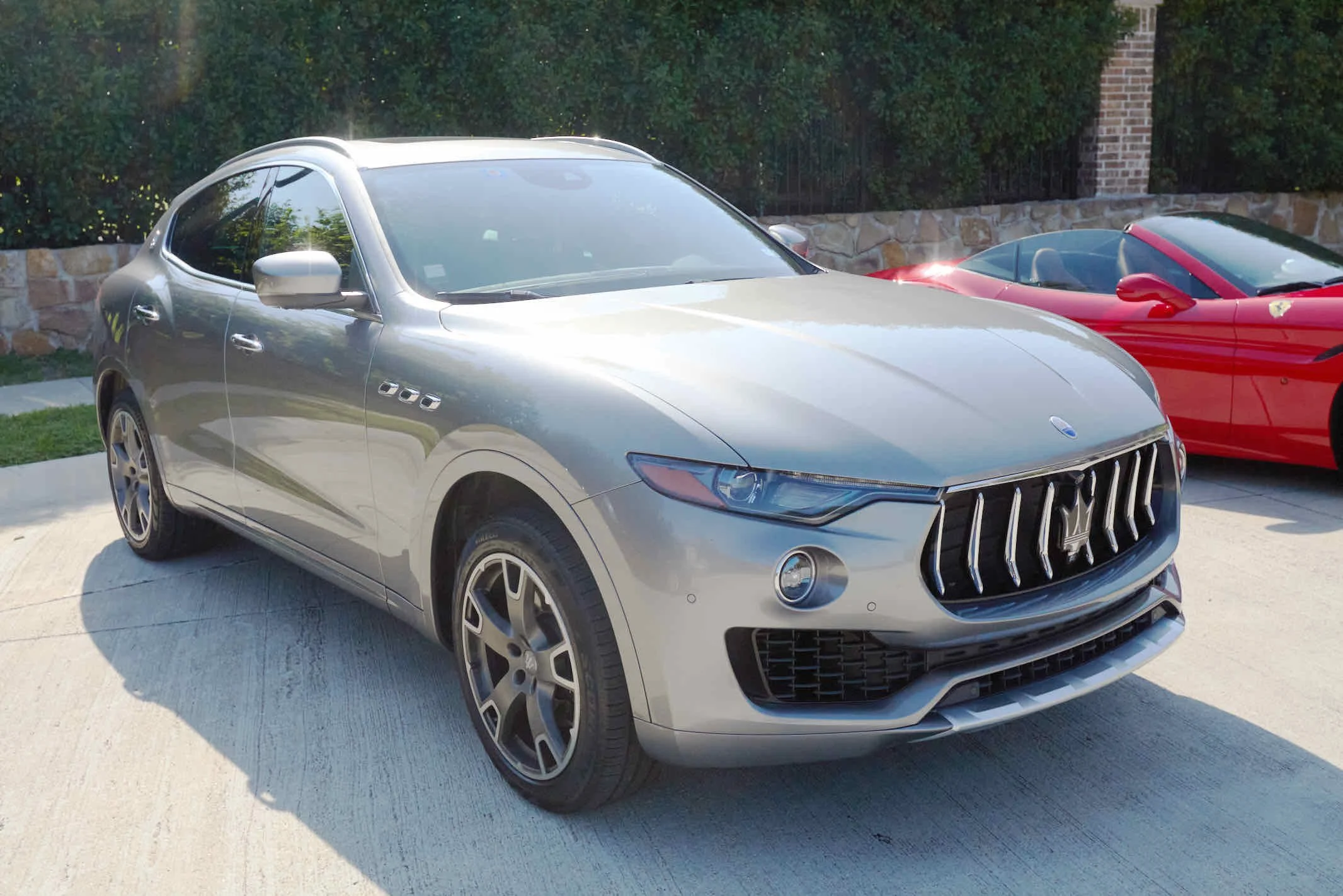 Silver luxury SUV parked on driveway with a red convertible sports car in background, greenery and stone wall behind.
