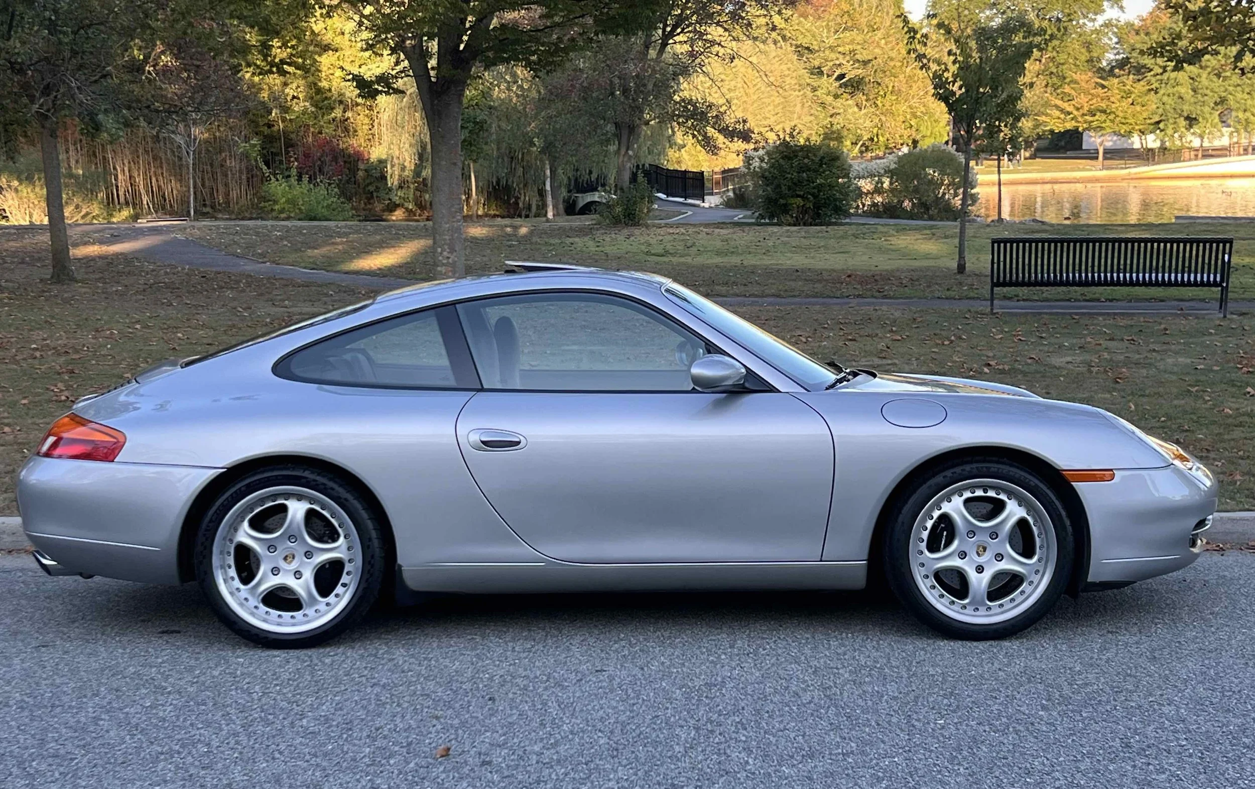 Side view of a silver Porsche 911 parked on the street with a park and lake in the background.