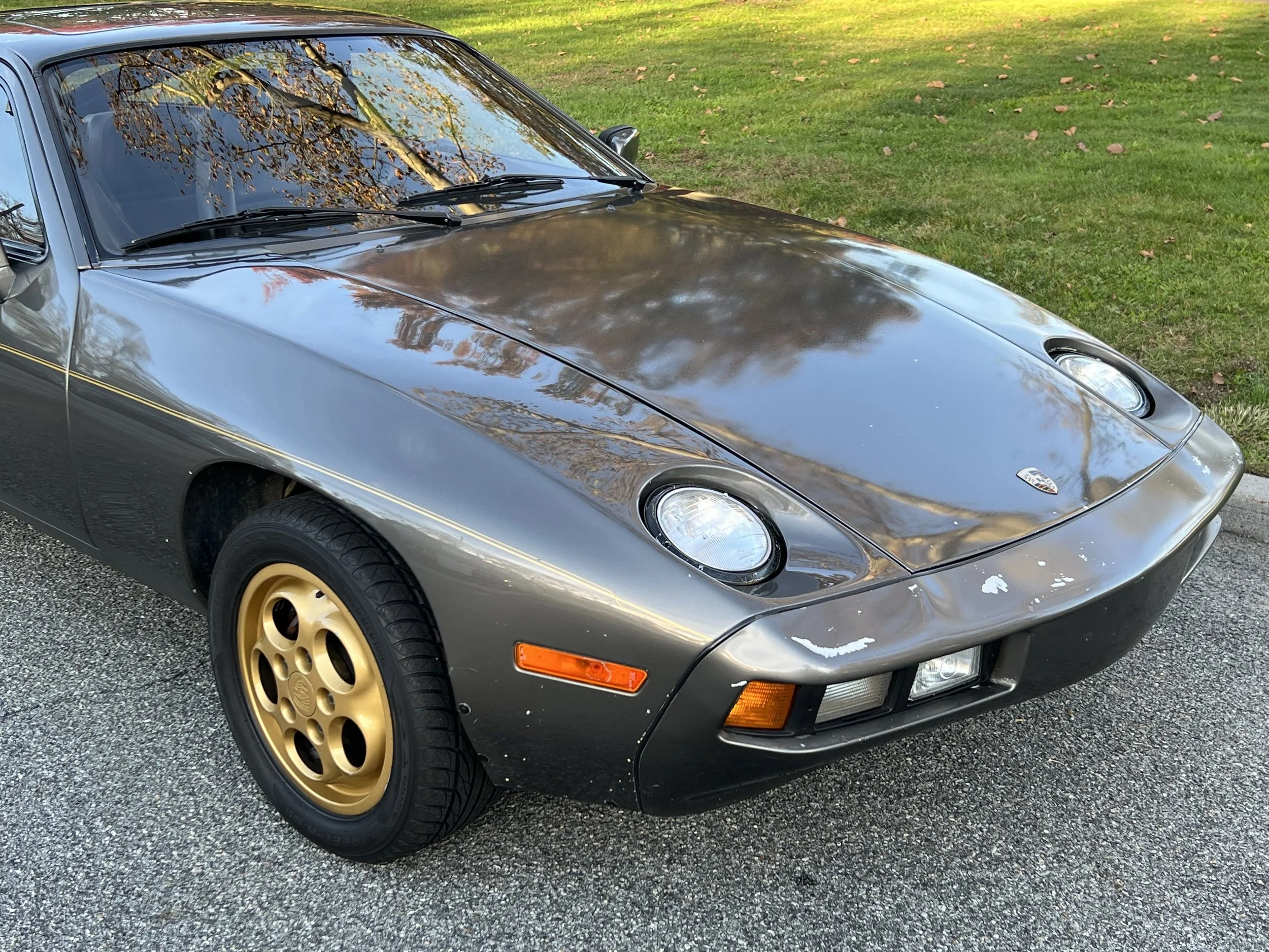 A vintage dark gray Porsche sports car with gold wheels parked on a driveway, reflecting trees and sky on its glossy surface.