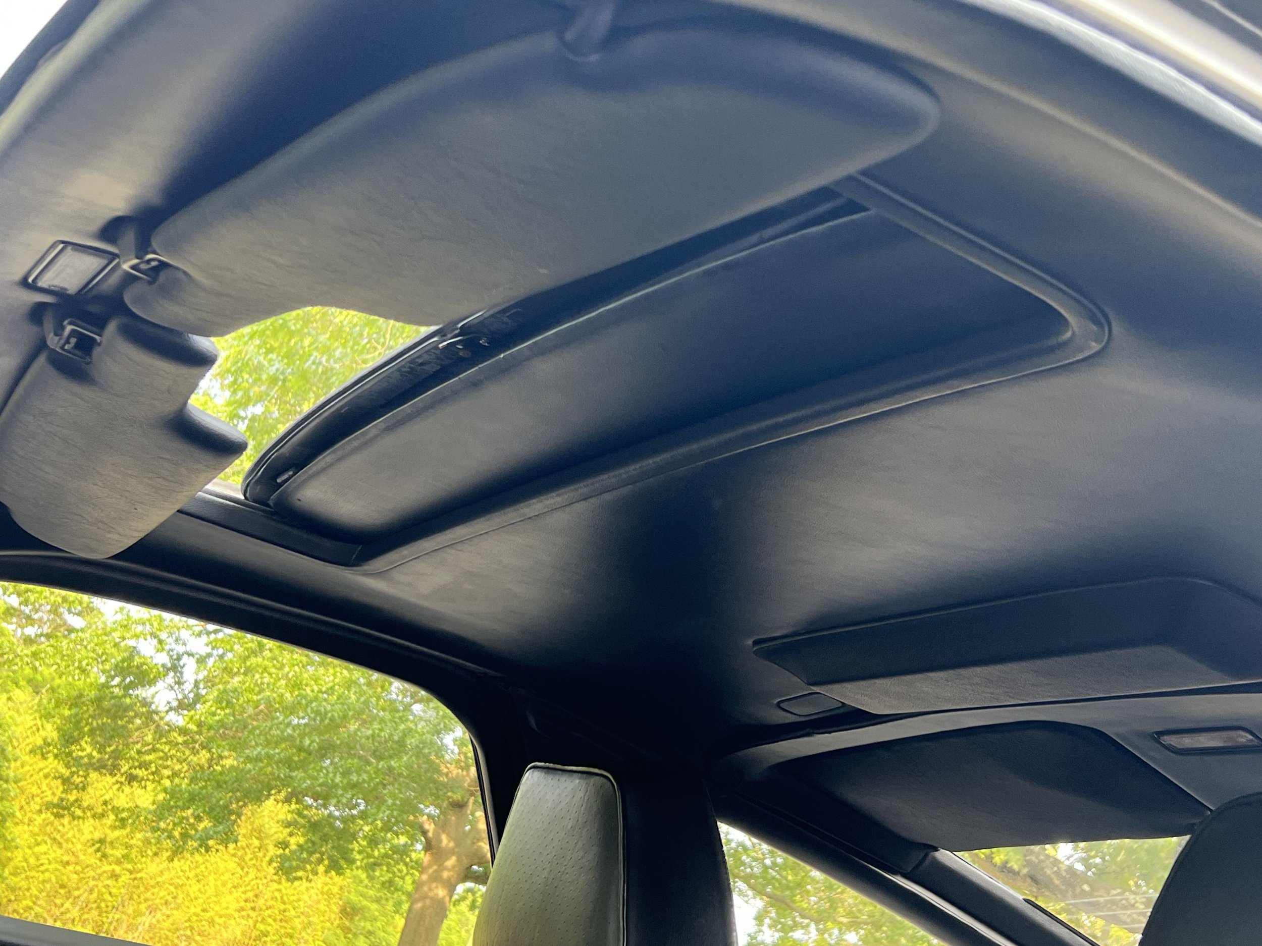 View of car's interior ceiling showing sunroof and overhead compartments with outside trees visible through the sunroof.