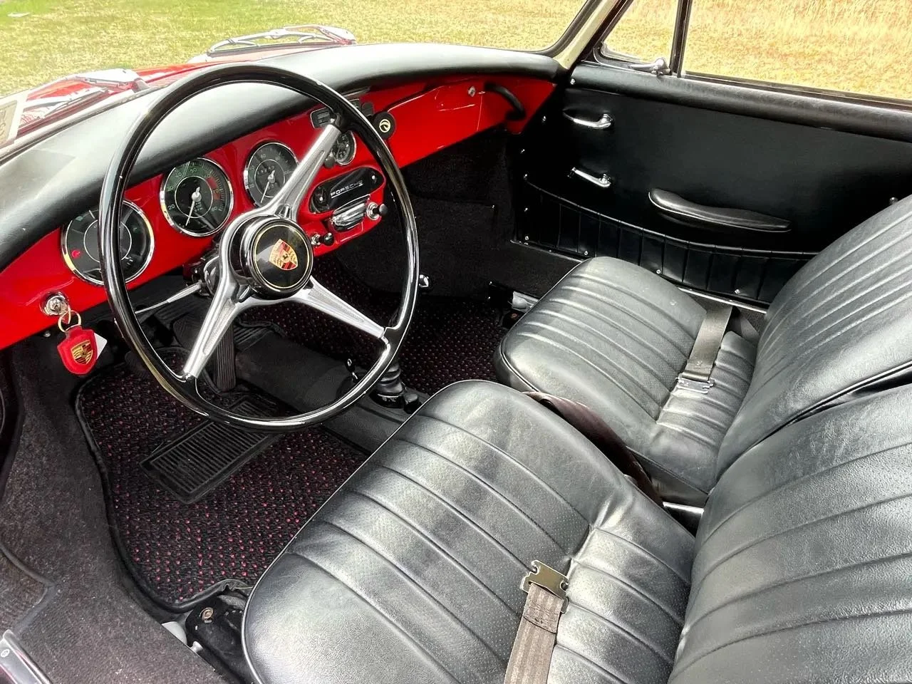 Interior of a vintage Porsche car featuring a black steering wheel with the Porsche logo, a red dashboard with multiple gauges, black leather front seats, and black door panels.