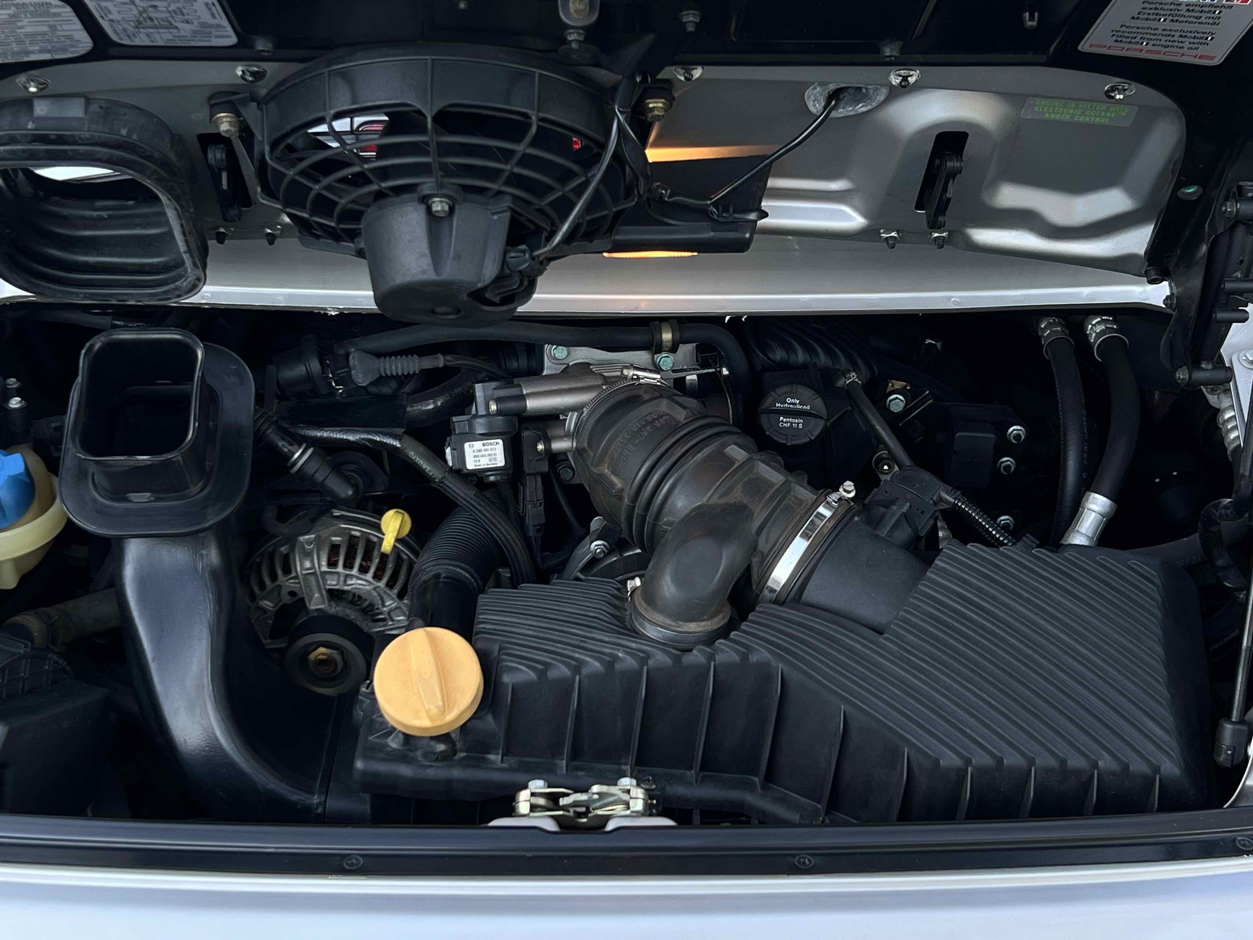 Car engine bay with visible air intake, alternator, and various hoses and components.