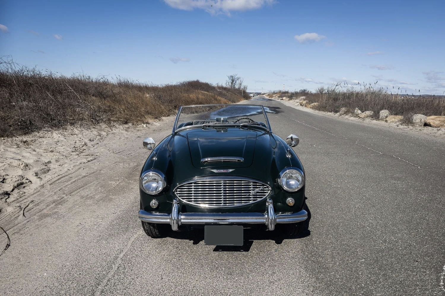 A vintage black convertible car parked on a deserted rural road with sparse bushes and rocks, under a blue sky with scattered clouds.