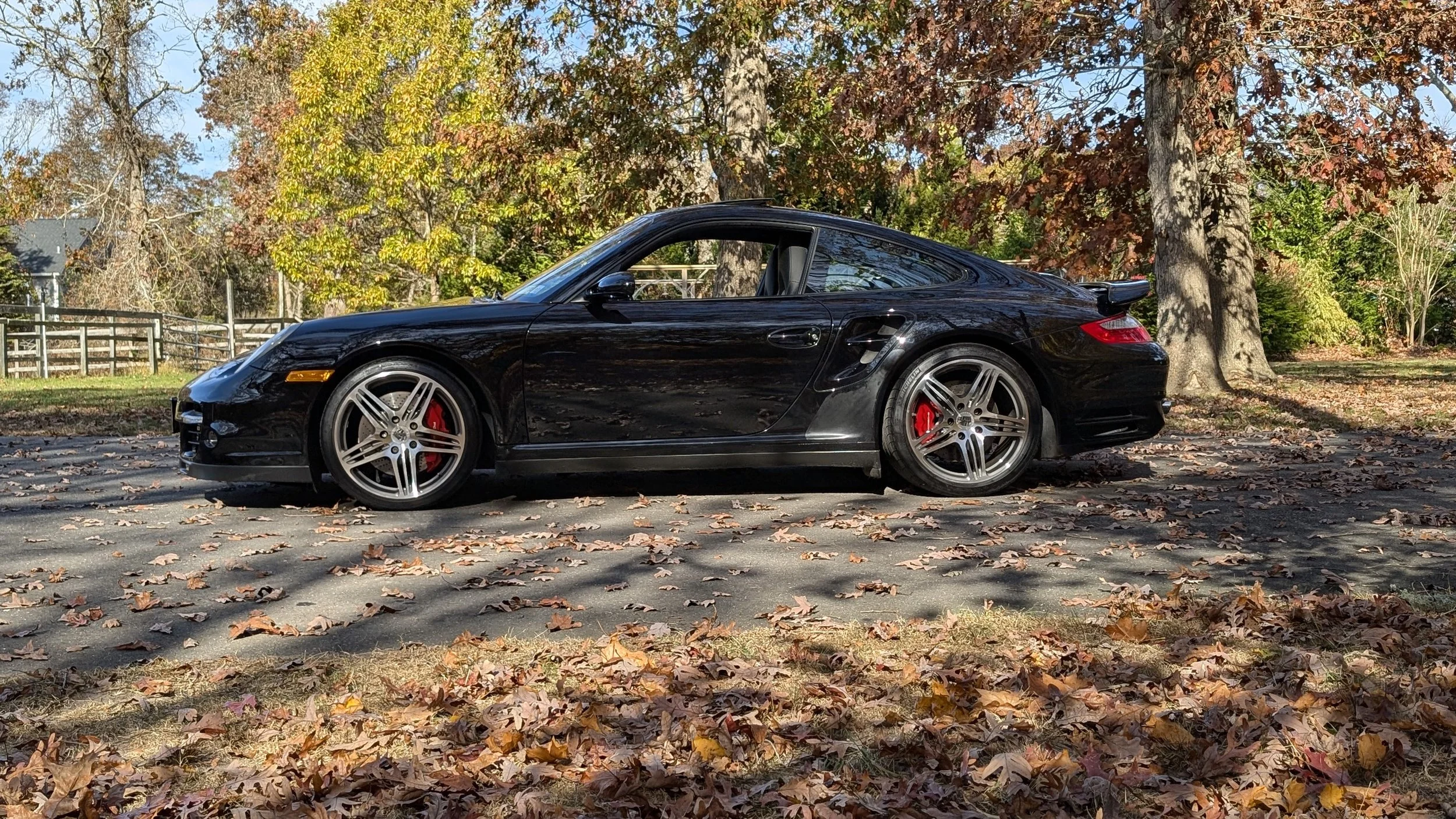 A black Porsche sports car parked on a paved area covered with fallen autumn leaves, with trees displaying colorful fall foliage in the background.