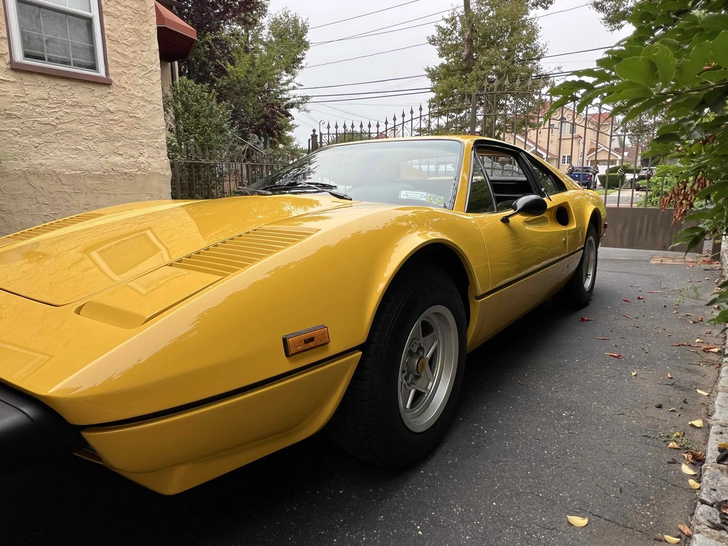 Yellow vintage sports car parked on a residential street with trees and a wrought iron gate in the background.
