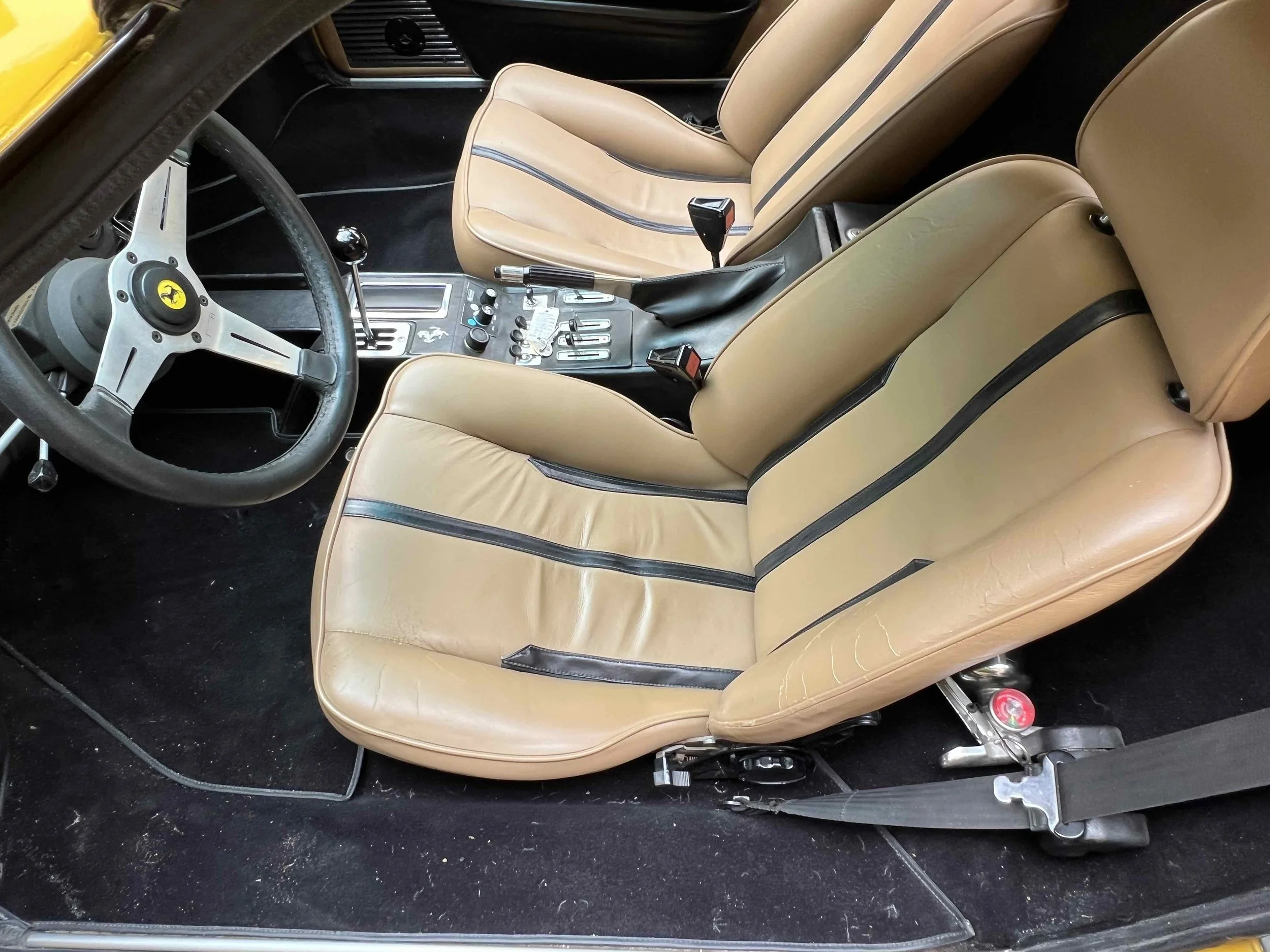 Interior of a vintage Ferrari with tan leather seats, black dashboard, and classic steering wheel, showing the driver and passenger seats, gear shifter, and control panel.