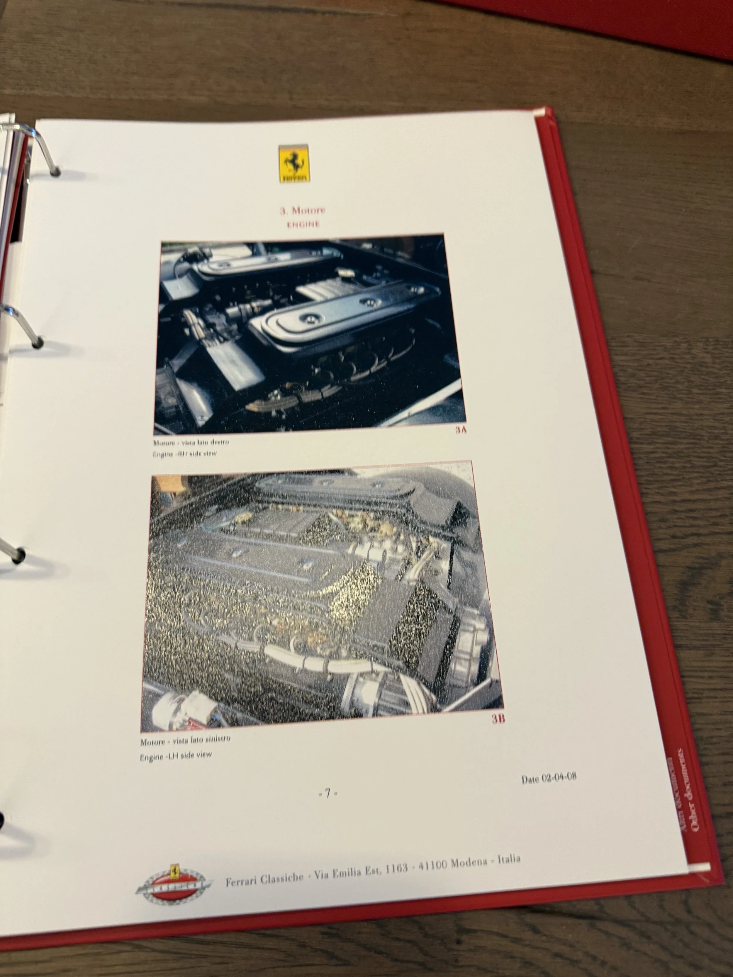 Photograph of a page from a manual showing two images of car engines, with Ferrari logo at the top and details at the bottom, on a wooden table.