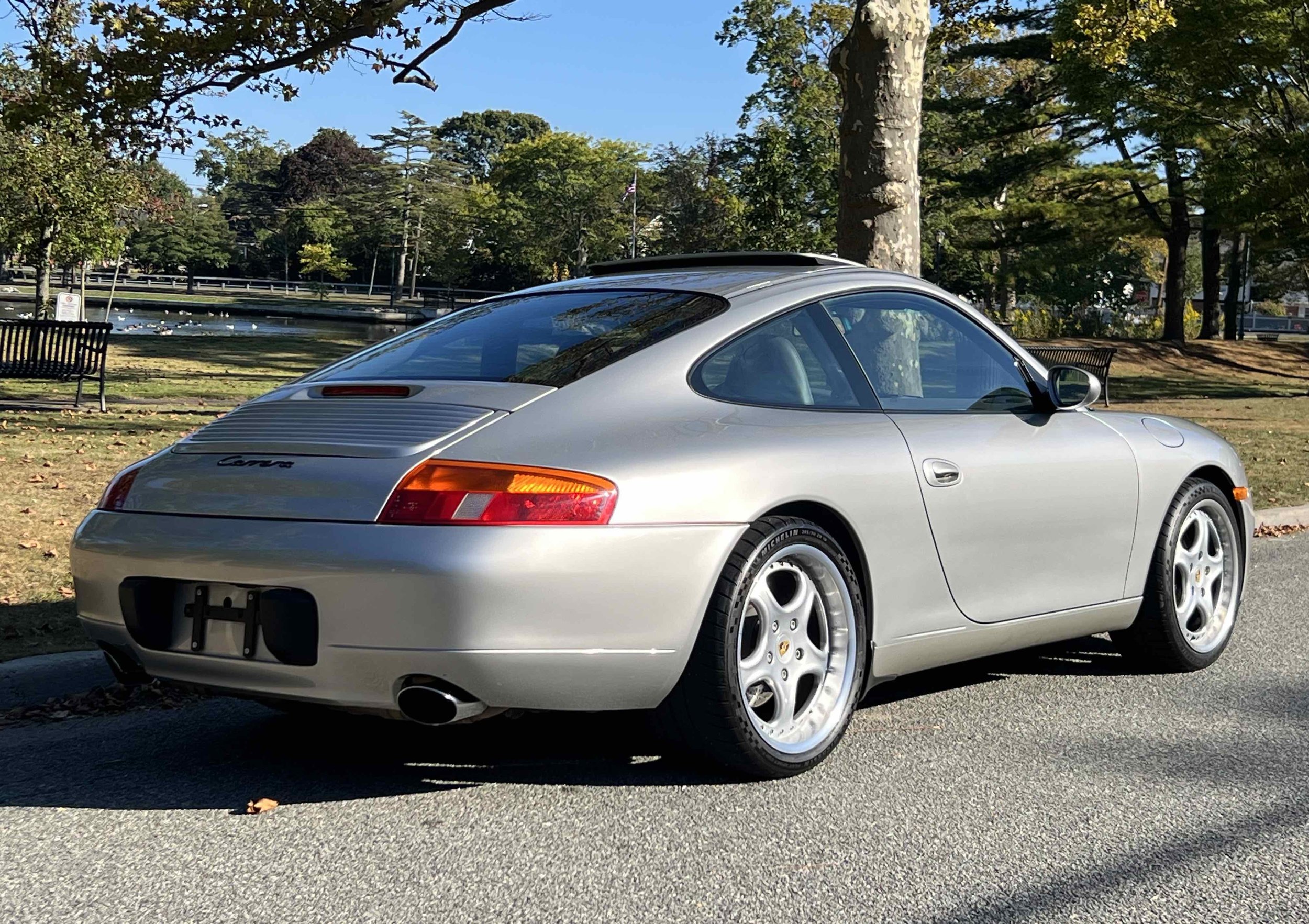 Silver Porsche 911 Carrera parked on street in park with trees and pond in background.