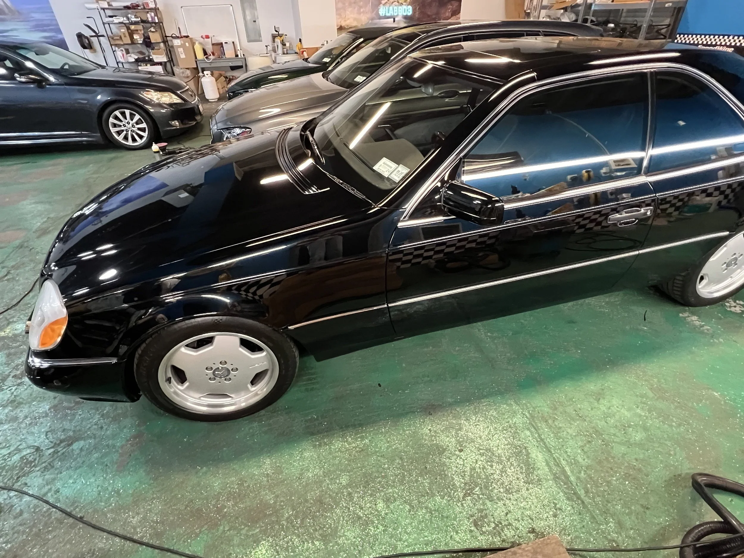 A black luxury Mercedes-Benz coupe parked indoors with other cars and a workshop or garage setting in the background.