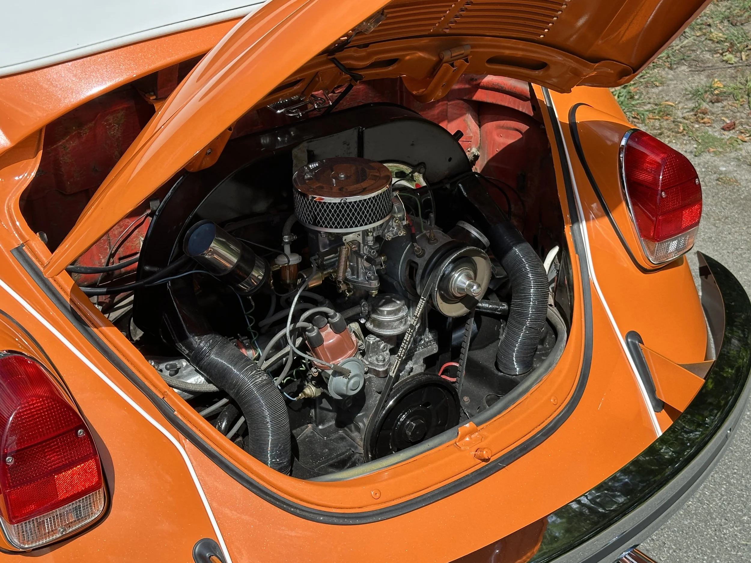 Open rear engine hatch of an orange classic car, showing the engine with chrome air filter and various mechanical parts.