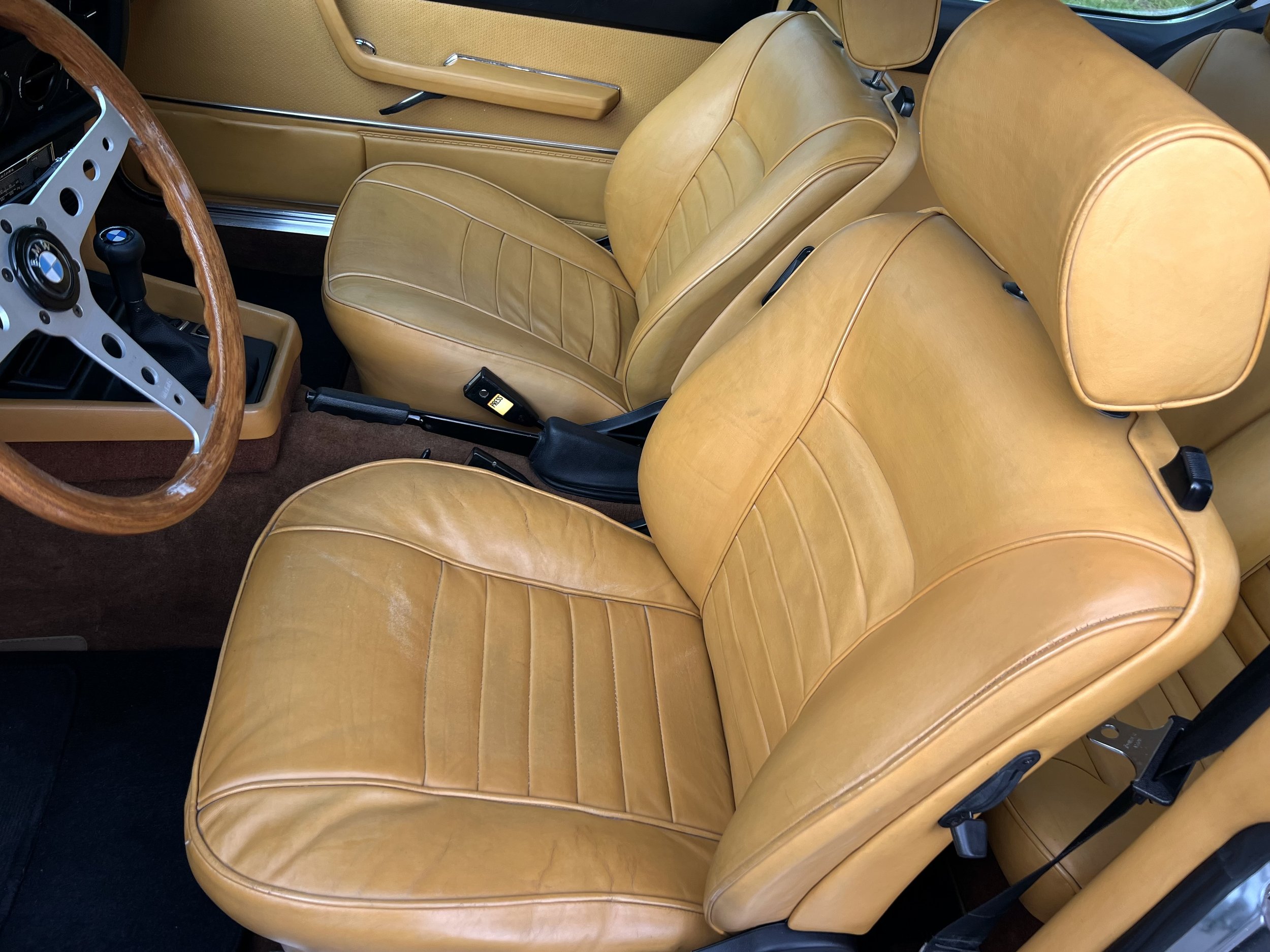 Interior of a vintage BMW 630 csi car showing tan leather seats, a steering wheel with a wooden rim, and a gear shift.