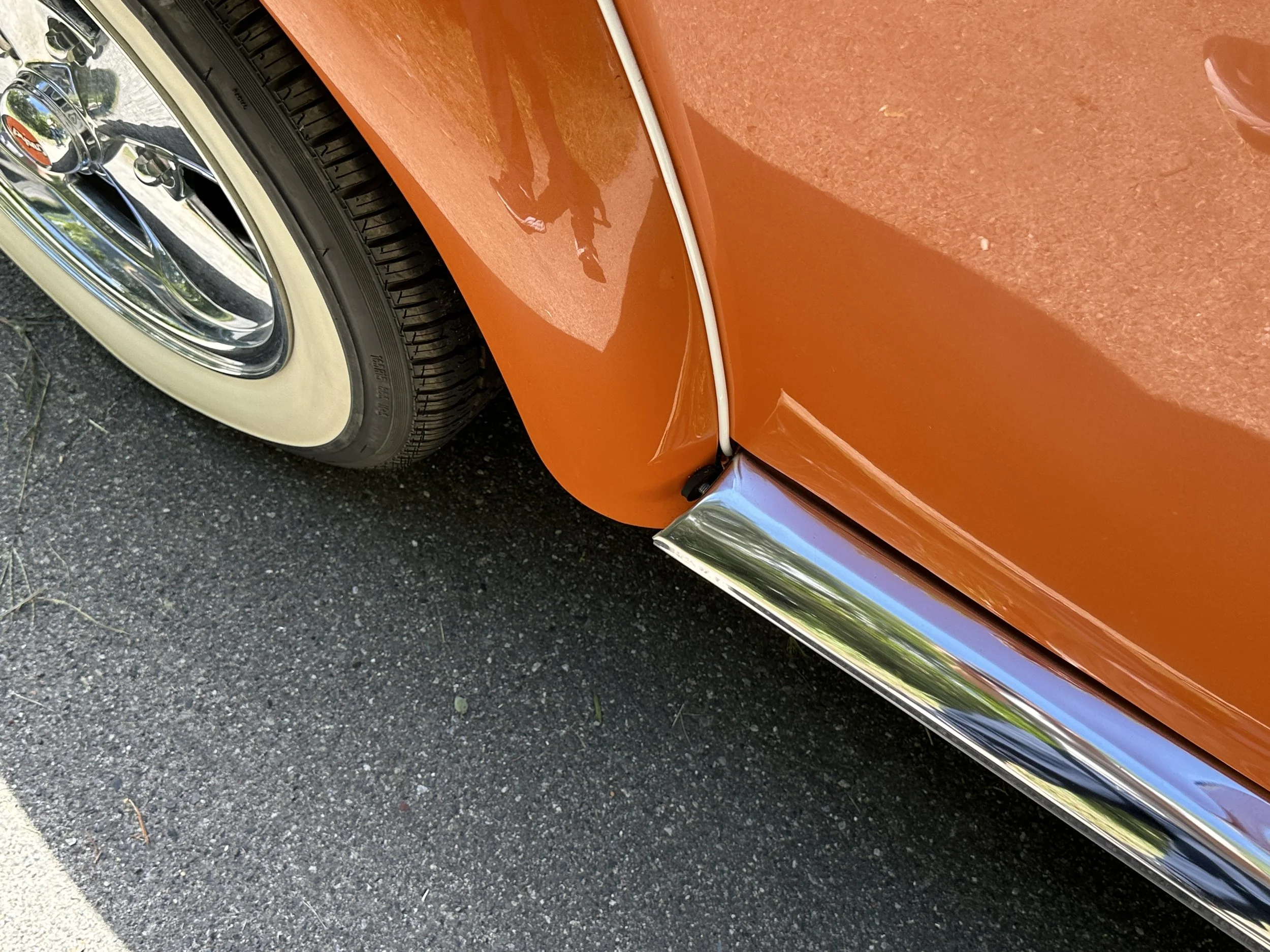 Close-up of a vintage orange car showing part of the wheel and fender, with a chrome trim running along the bottom edge.