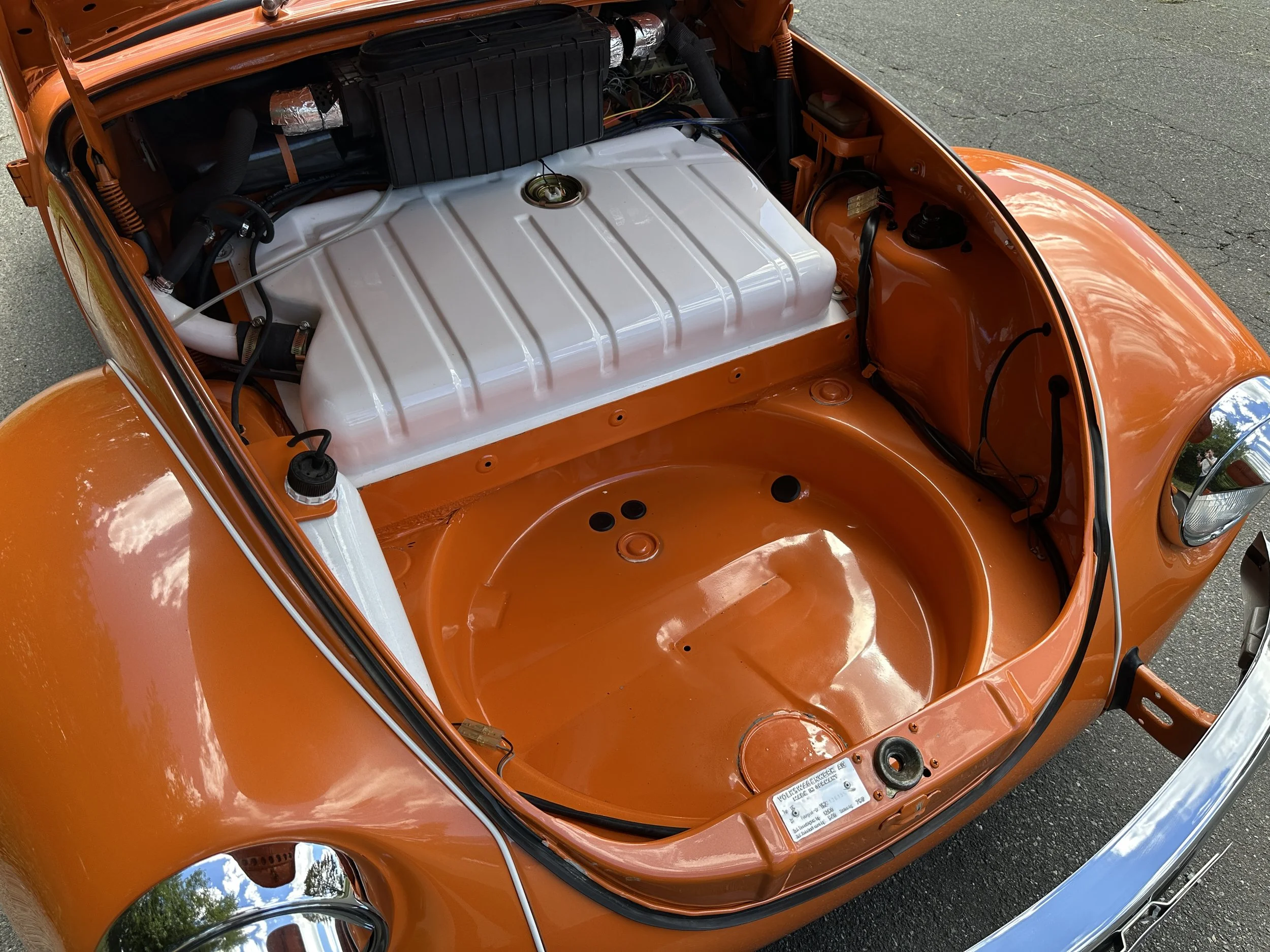 The front trunk of an orange vintage Volkswagen Beetle with no engine, showing the empty space, fuel tank, and various internal components.