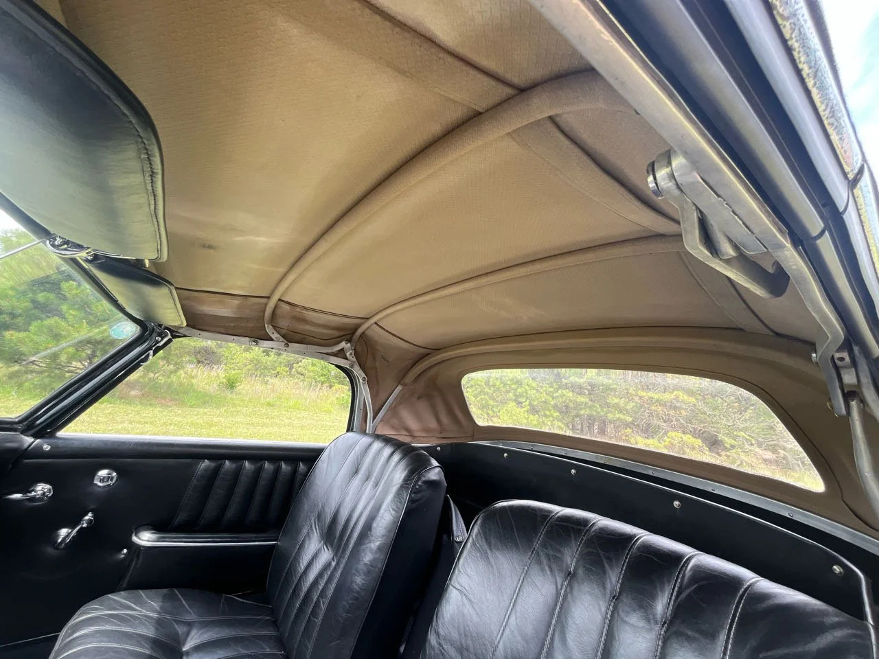 Interior of a vintage car showing black leather seat, door panel with window crank and handle, and beige fabric ceiling with support beams.