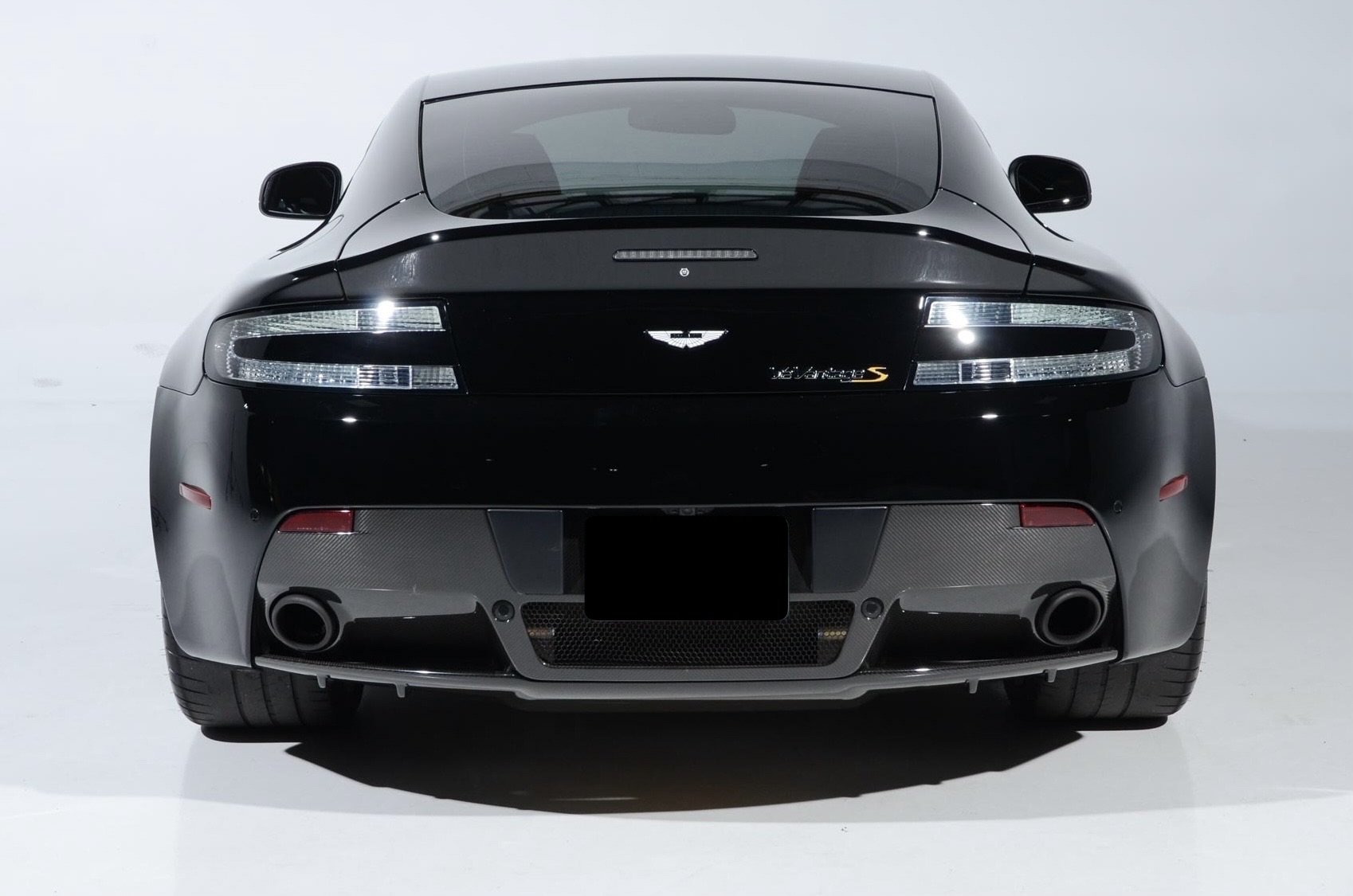 Rear view of a black Aston Martin V8 Vantage S sports car with LED taillights and a dual exhaust system on both sides, set against a plain white background.