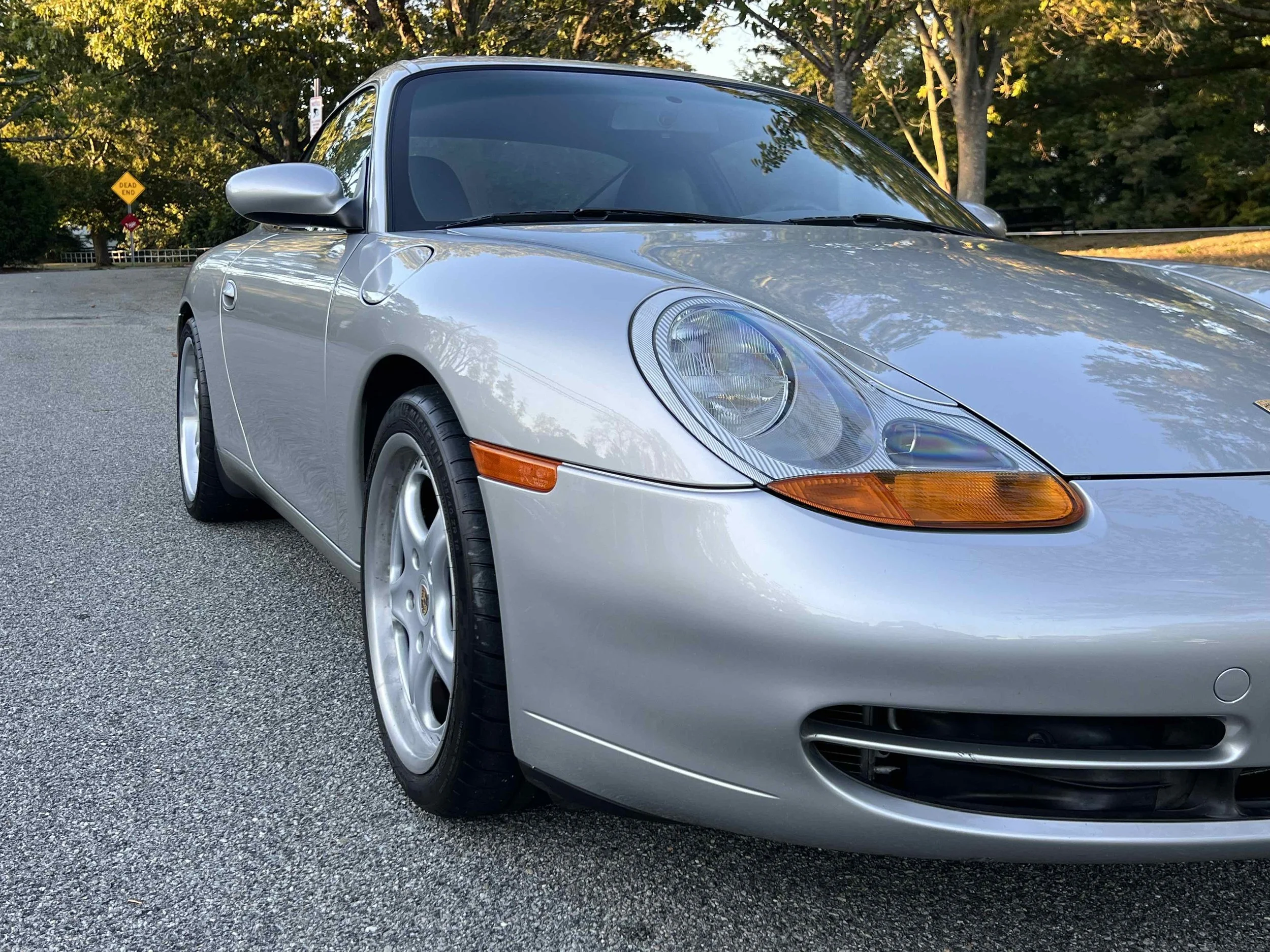 A silver sports car parked on a paved surface with trees and a yellow road sign in the background.