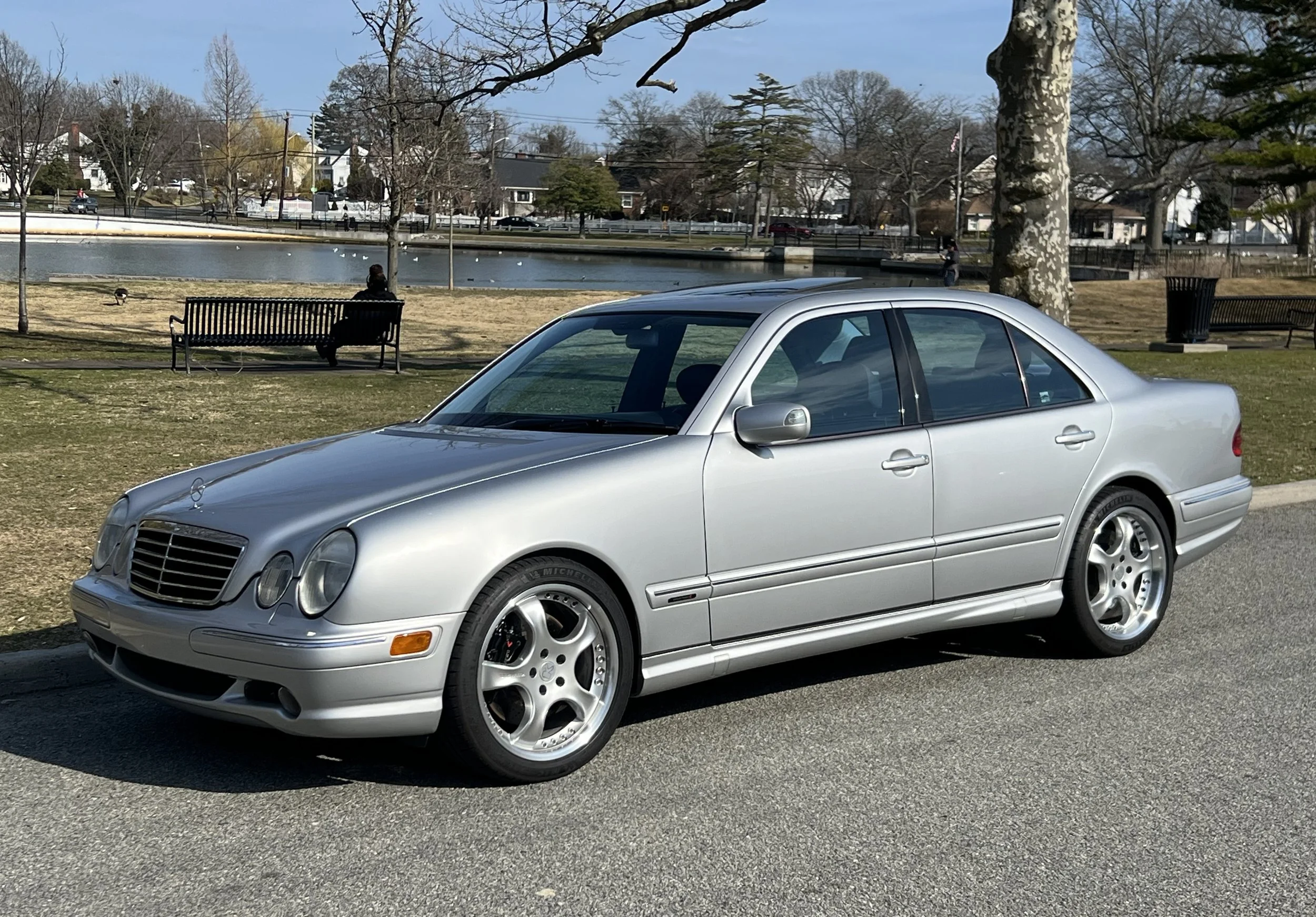 Silver Mercedes-Benz parked on a street near a park with benches, trees, and a pond in the background.
