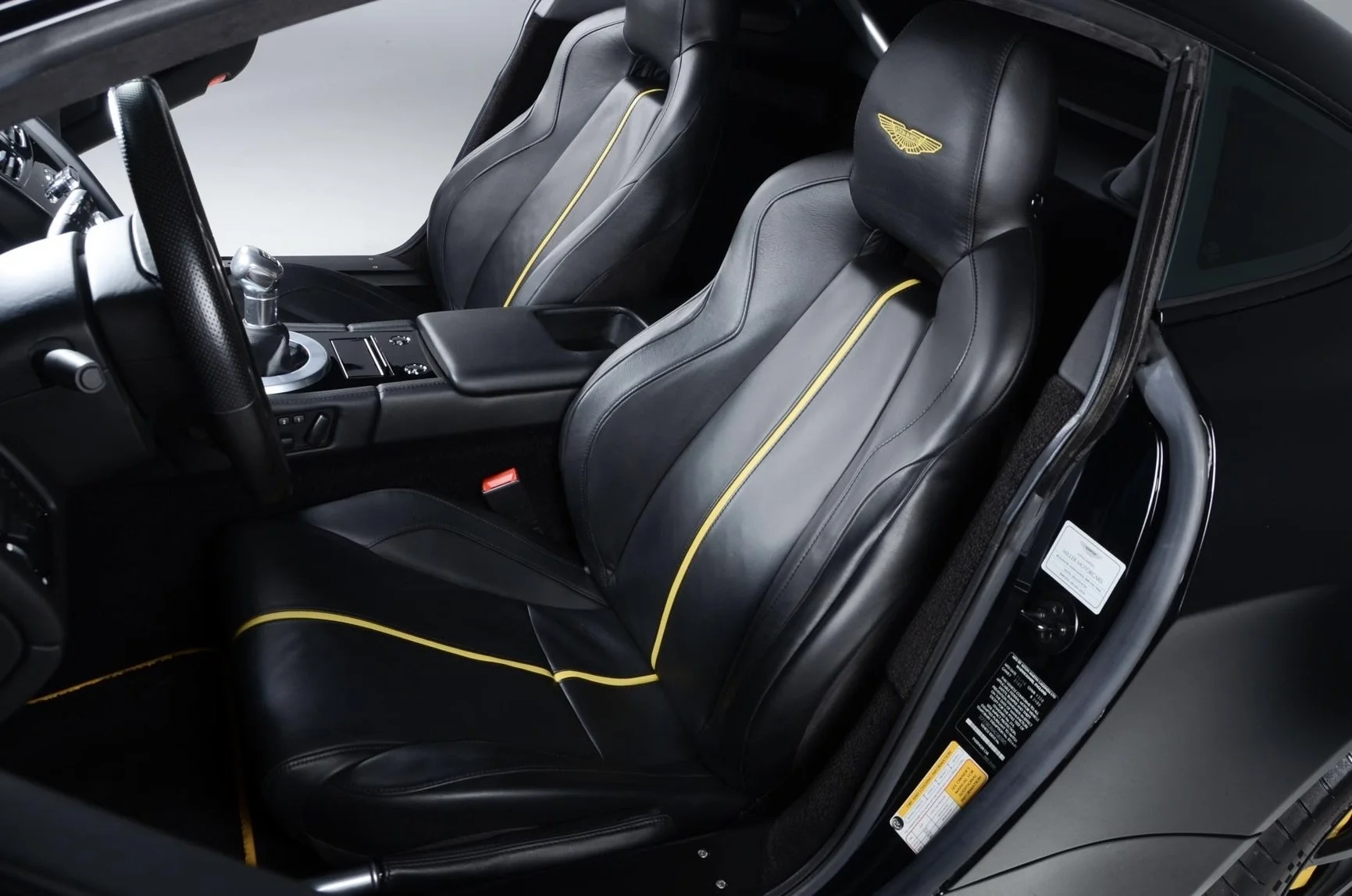 Interior of a luxury car with black leather seats featuring yellow piping and an embroidered logo on the headrests, with a gear shift, center console, and side door visible.