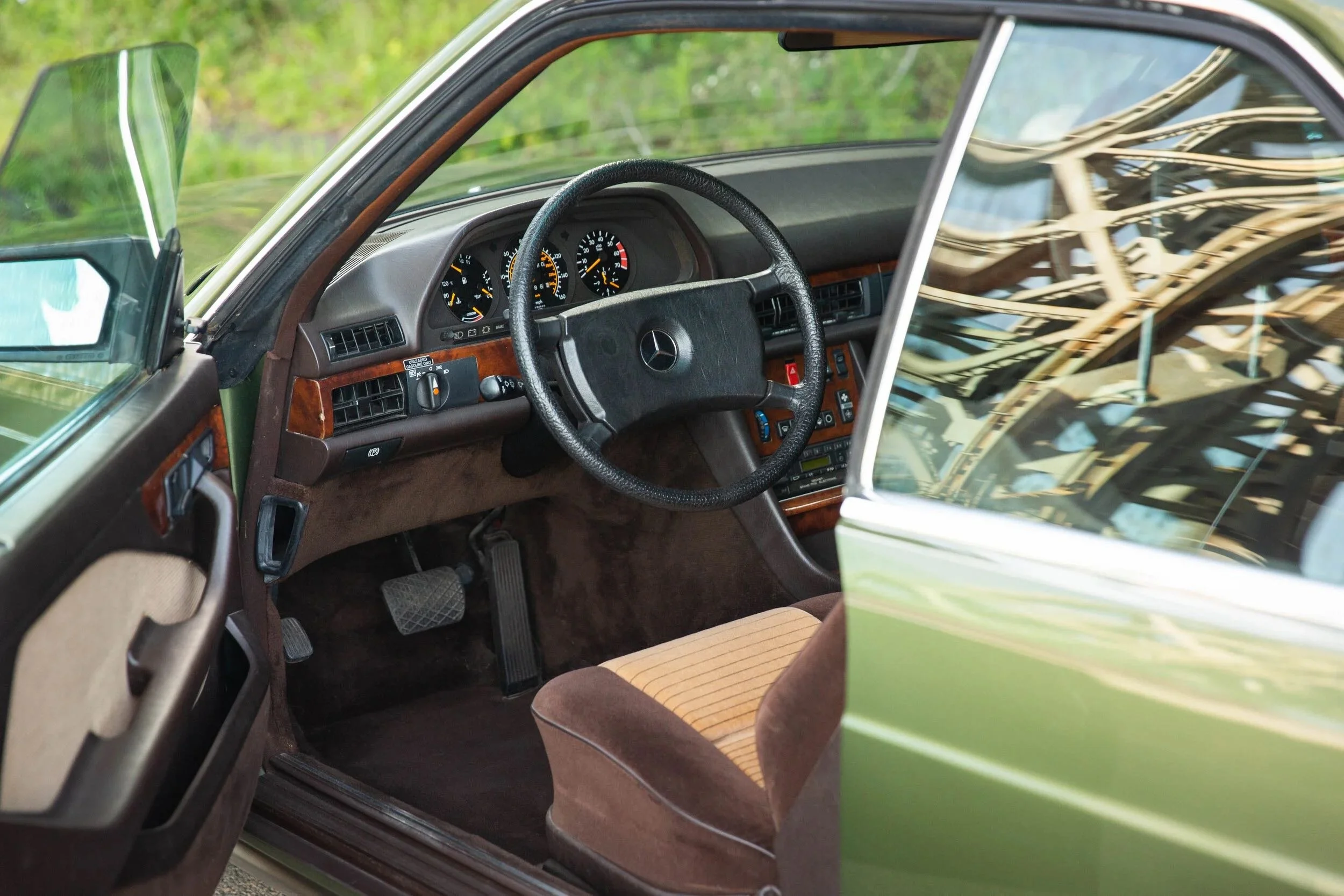 Interior of a vintage Mercedes-Benz car showing steering wheel, dashboard with gauges, and a front seat with brown upholstery, with the driver's side door open.