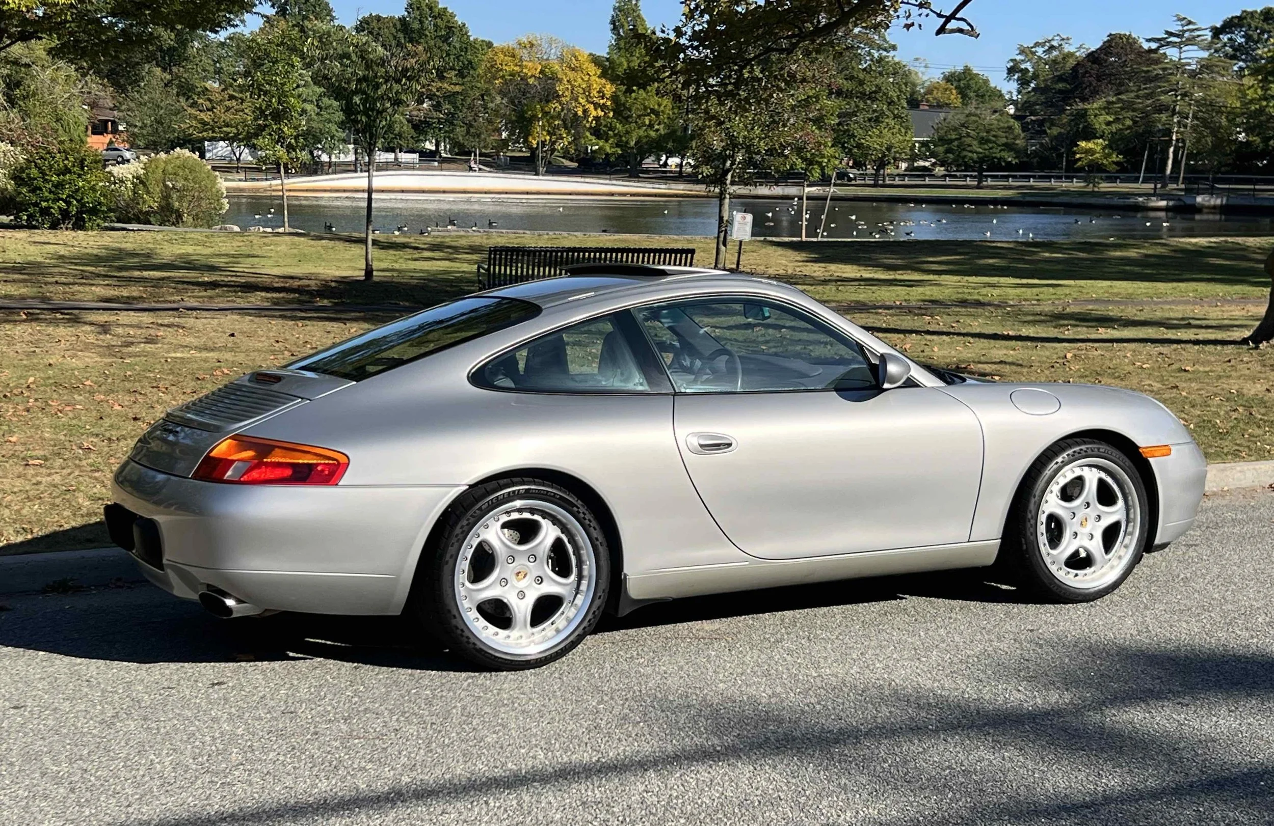 Silver Porsche 911 parked on the side of a street next to a grassy park with a pond and ducks, trees with fall foliage in the background on a sunny day.