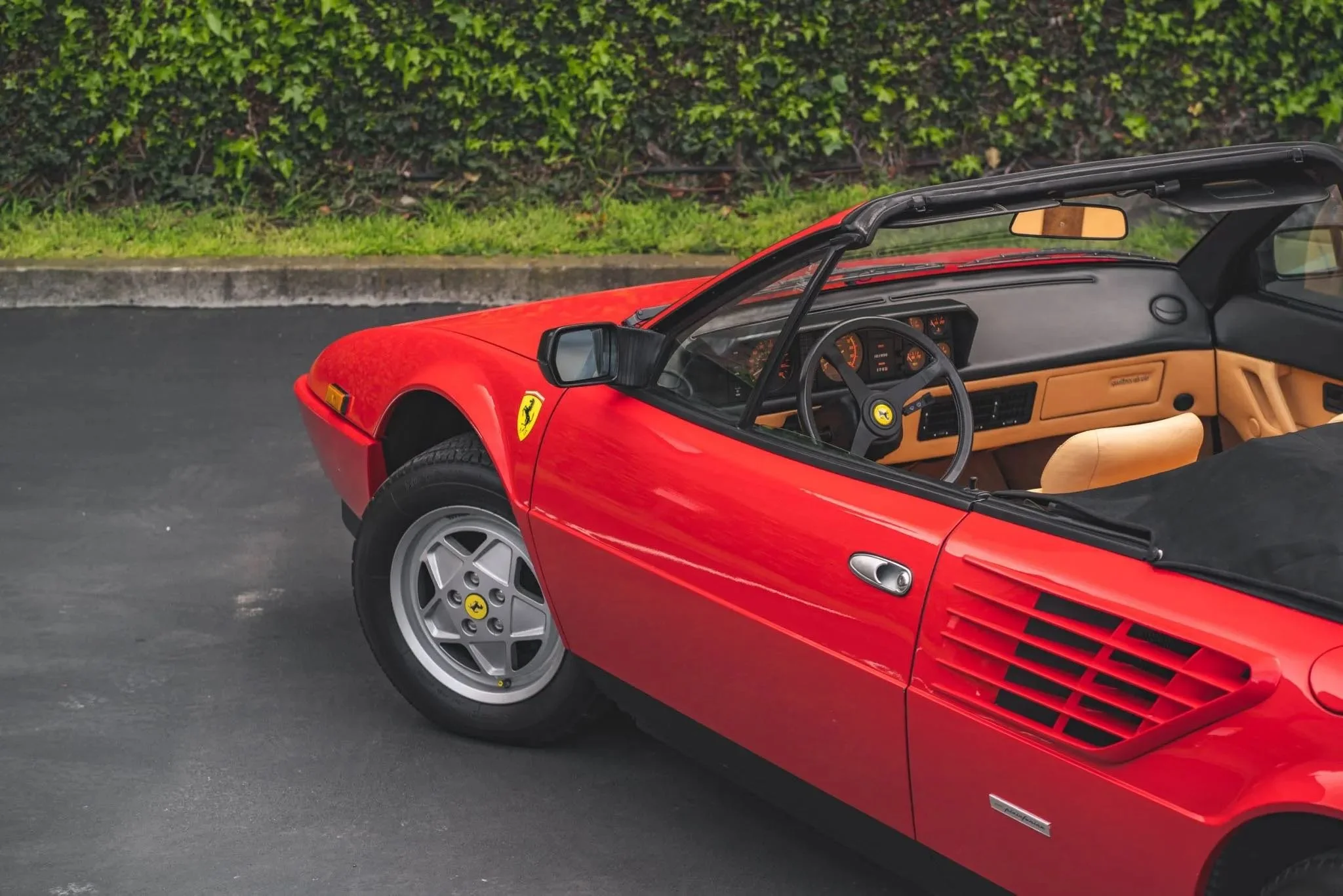 Red vintage Ferrari sports car parked on a dark asphalt surface with green hedge in the background.