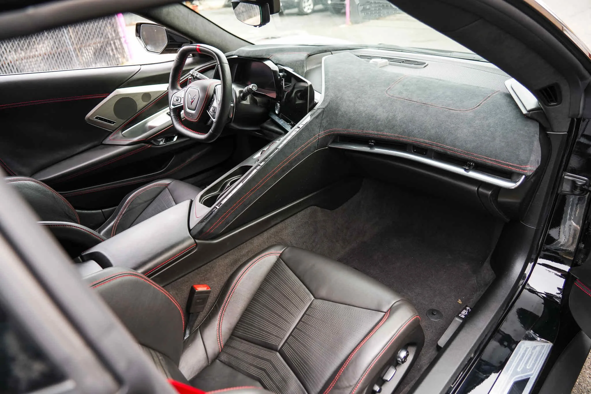 Interior of a sports car showing the driver's seat, dashboard, and center console with black leather seats with red stitching.