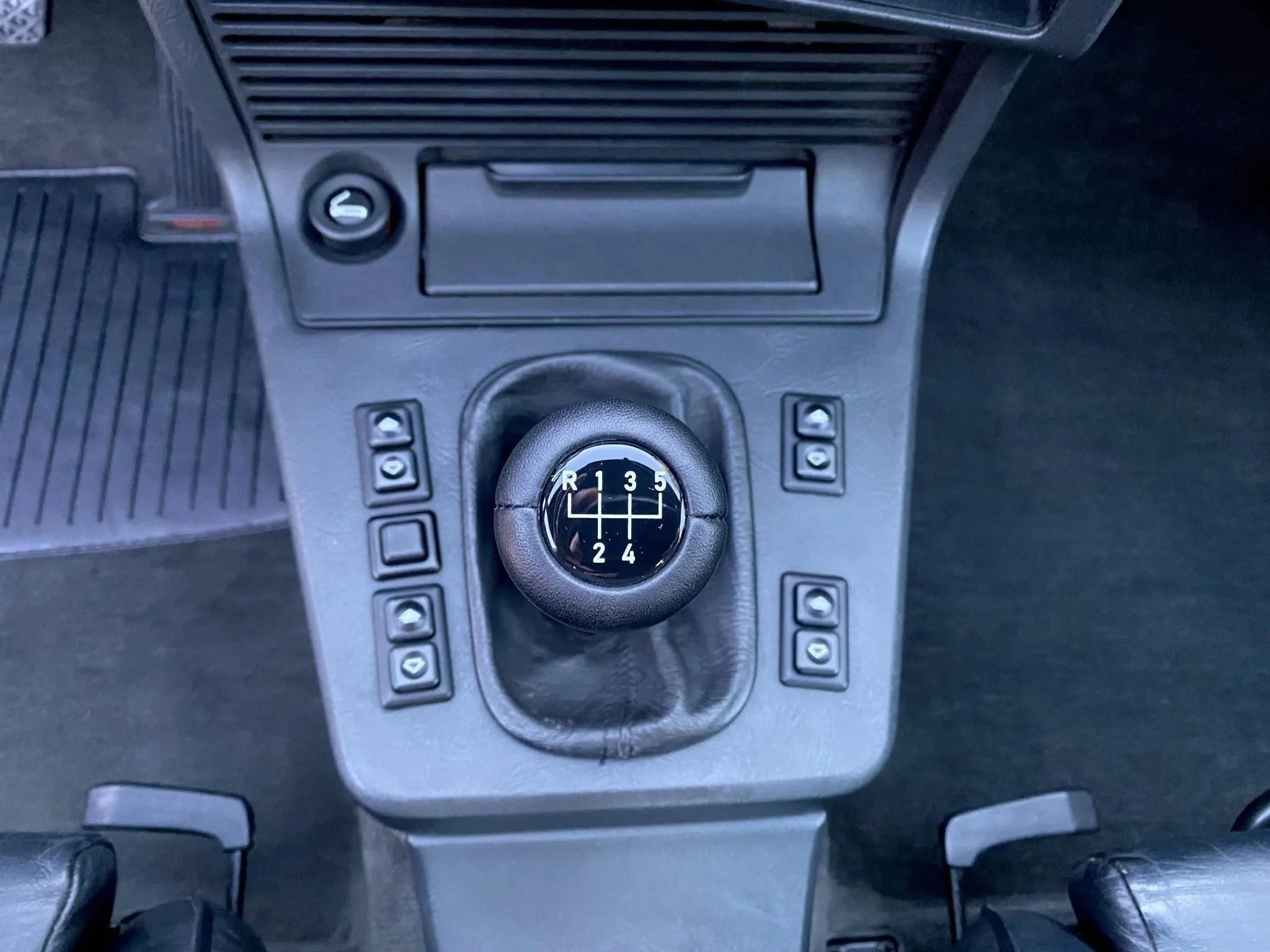 Top view of a manual gear shift in a car with six gears plus reverse, surrounded by control buttons.