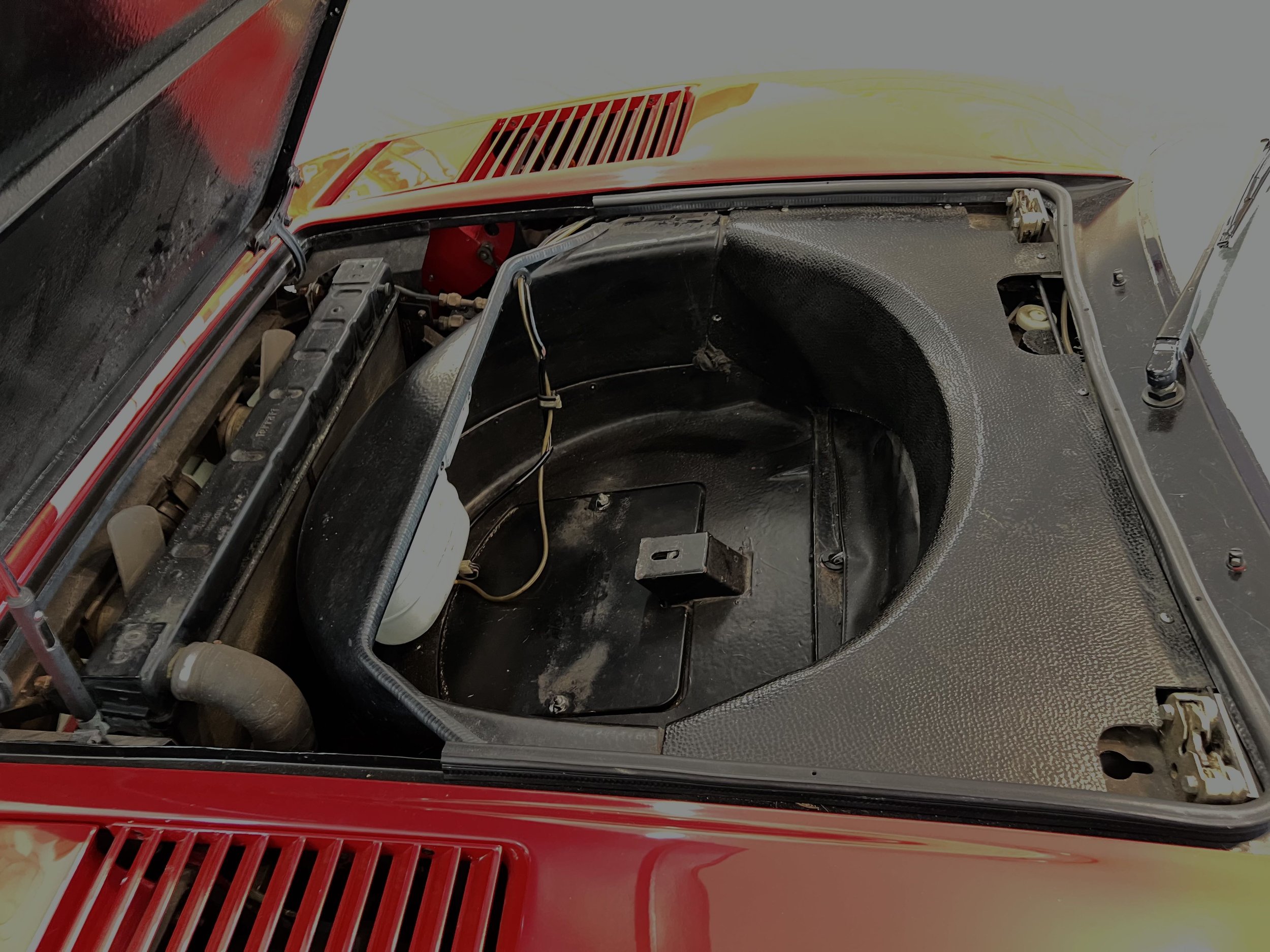 The front trunk of a red vintage sports car with the hood open, showing the empty storage compartment and some exposed components.