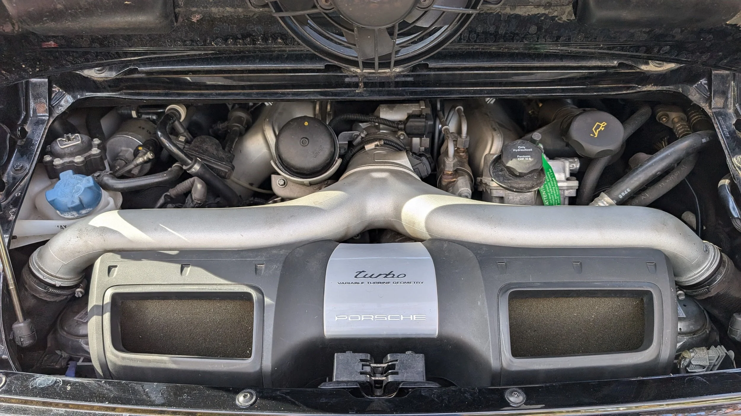 The image shows the engine bay of a Porsche sports car, featuring a turbocharged engine with visible components such as intake manifolds and various engine hoses. The engine cover prominently displays the Porsche logo and a label indicating it is a t