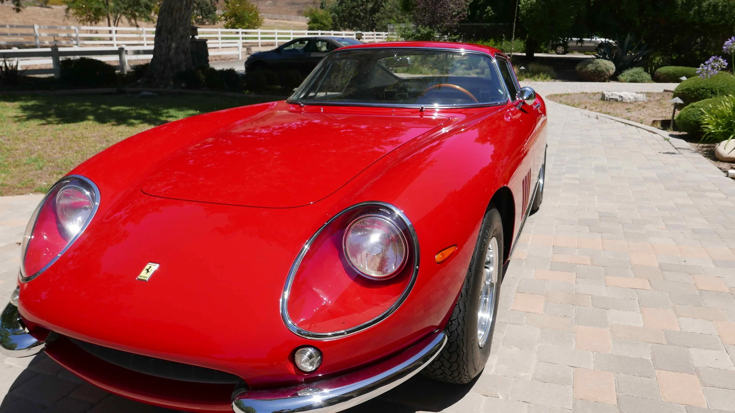 Red vintage Ferrari sports car parked on a paved driveway with green shrubs, trees, and a white fence in the background.