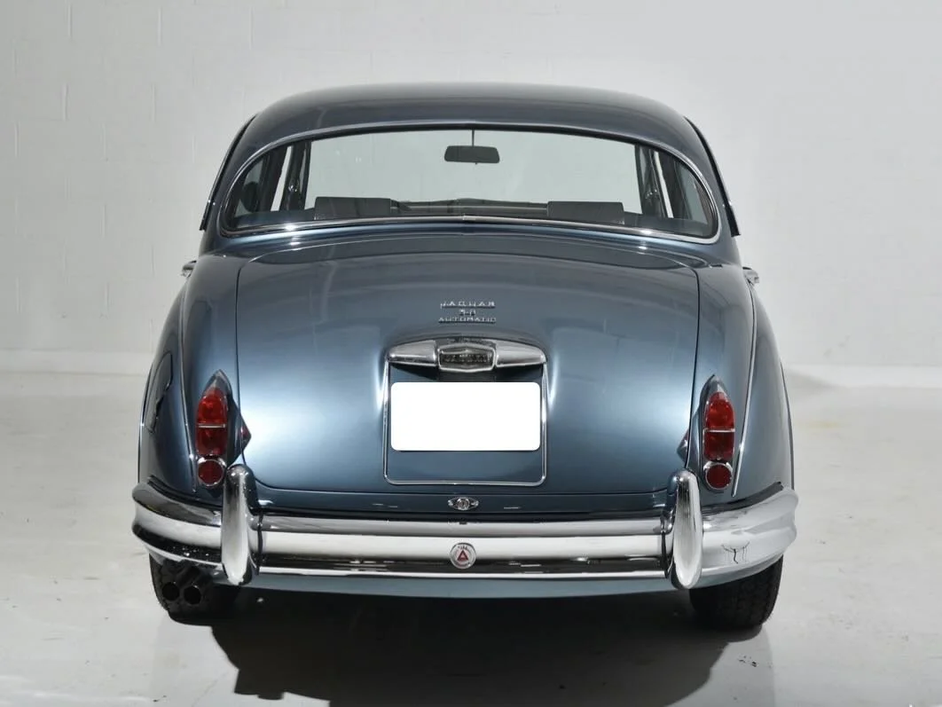 Rear view of a vintage blue Jaguar automobile with chrome bumpers and tail lights in a plain, well-lit room.
