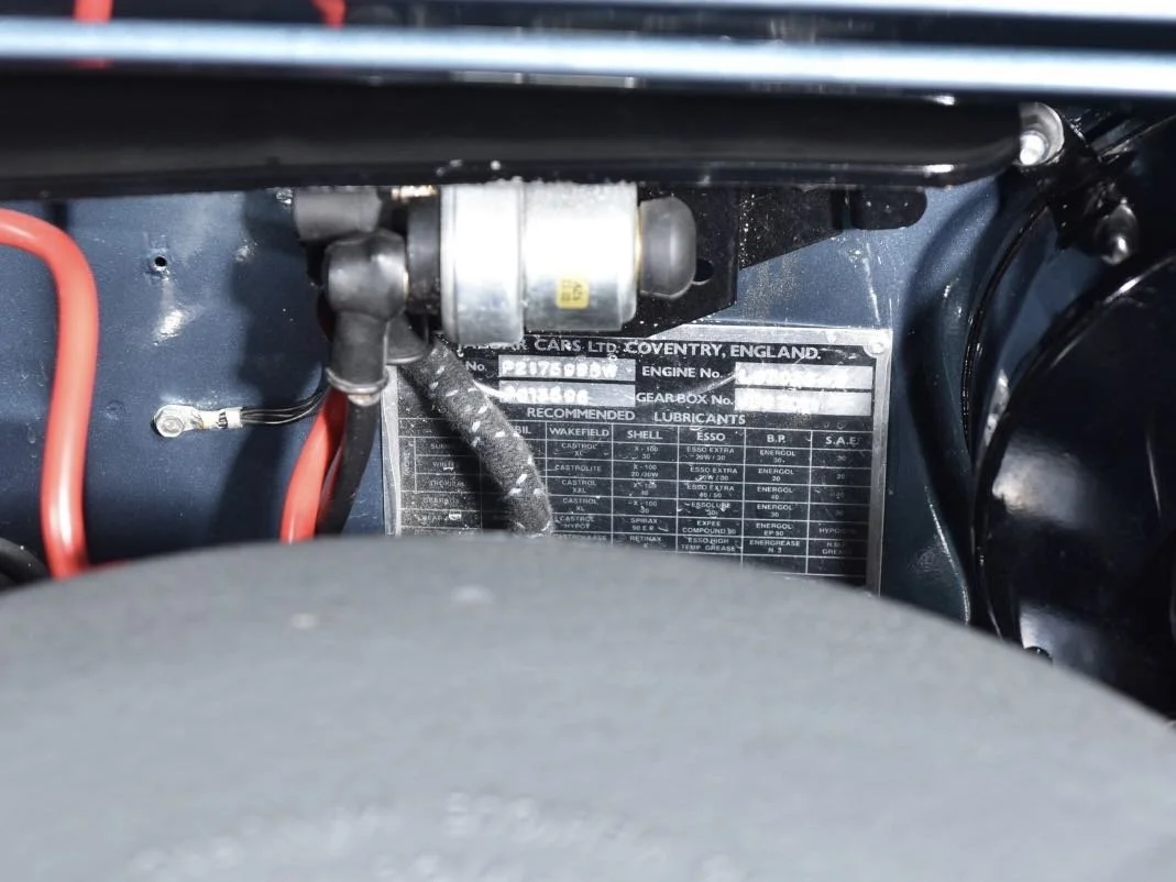 Close-up of a vintage car engine compartment with a metal information plate showing manufacturer details, engine number, gearbox number, and recommended lubricants, surrounded by various engine components and wiring.