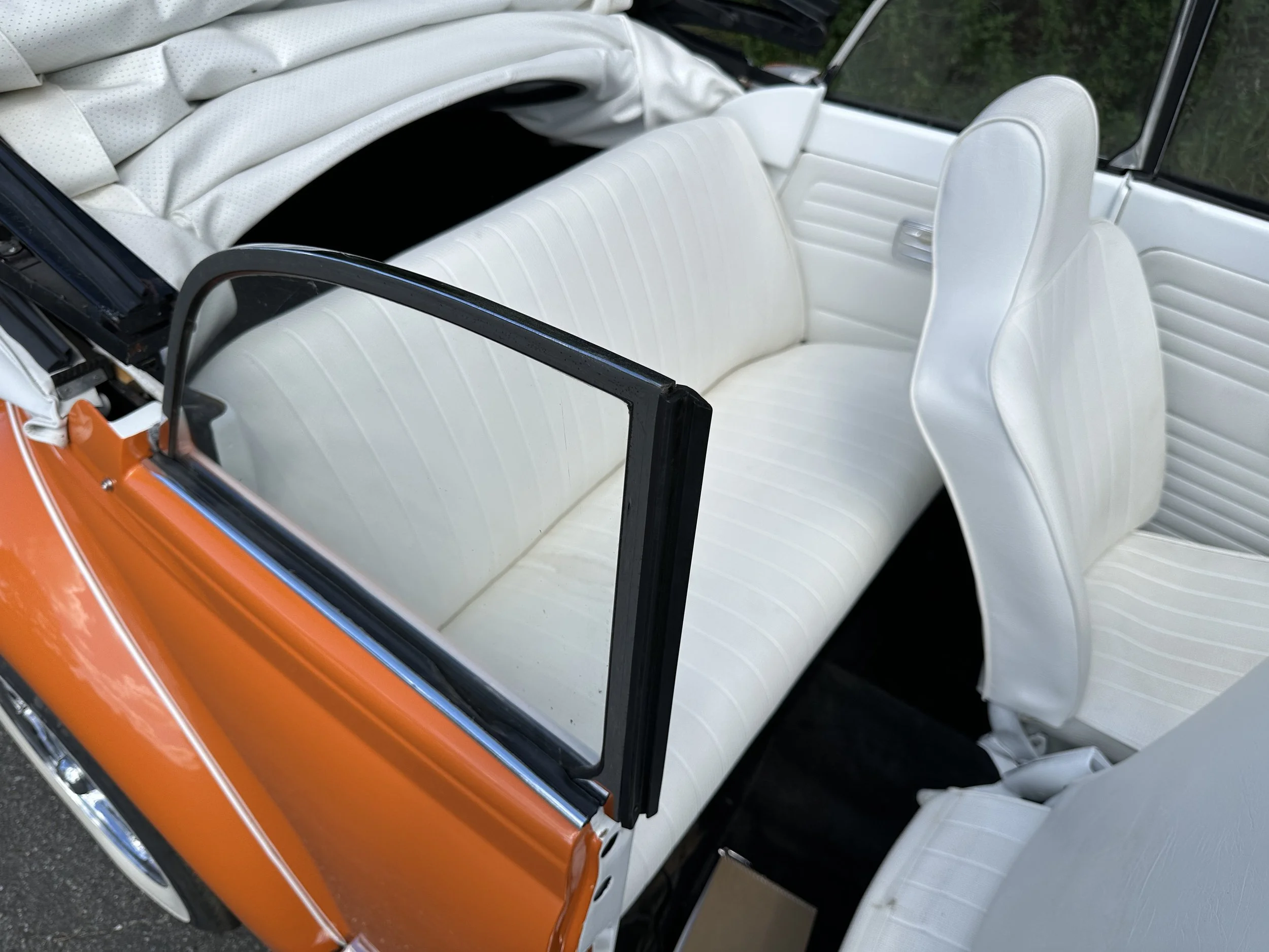 The image shows the interior of a vintage car with white upholstered seats, with some parts of the car's exterior being orange. The car appears to be a classic model with a fold-down roof and open top.