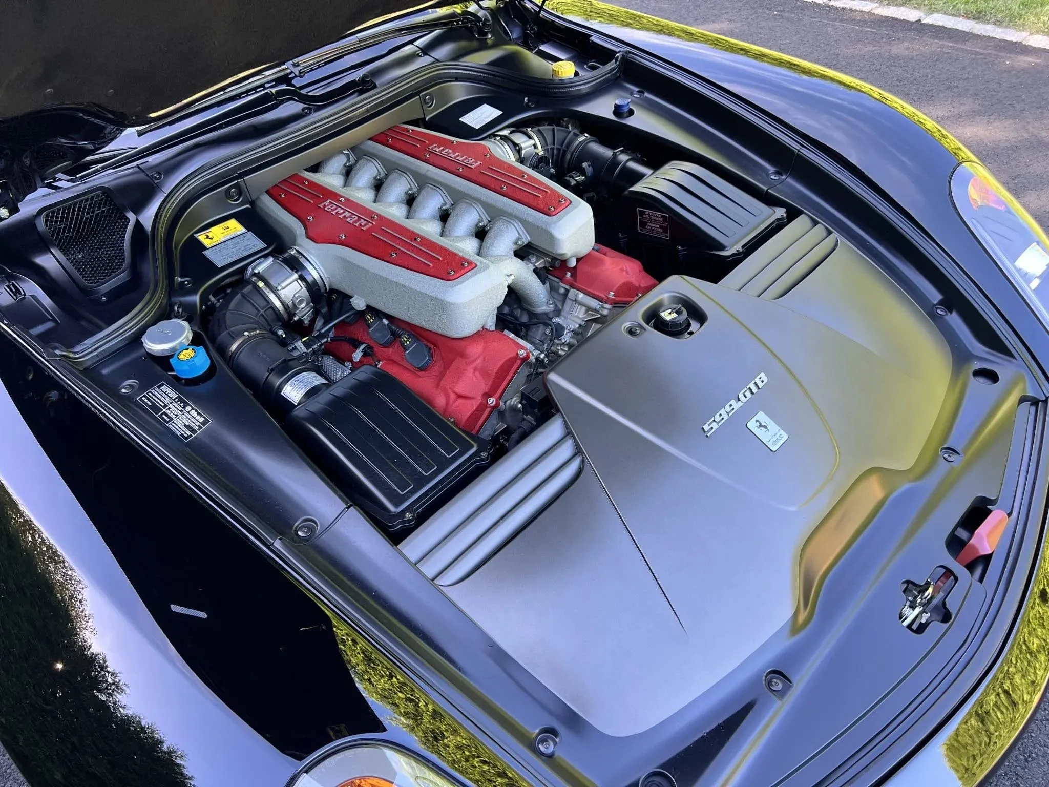 Close-up view of a Ferrari sports car engine under the hood, showing silver and red engine components, with the Ferrari logo.