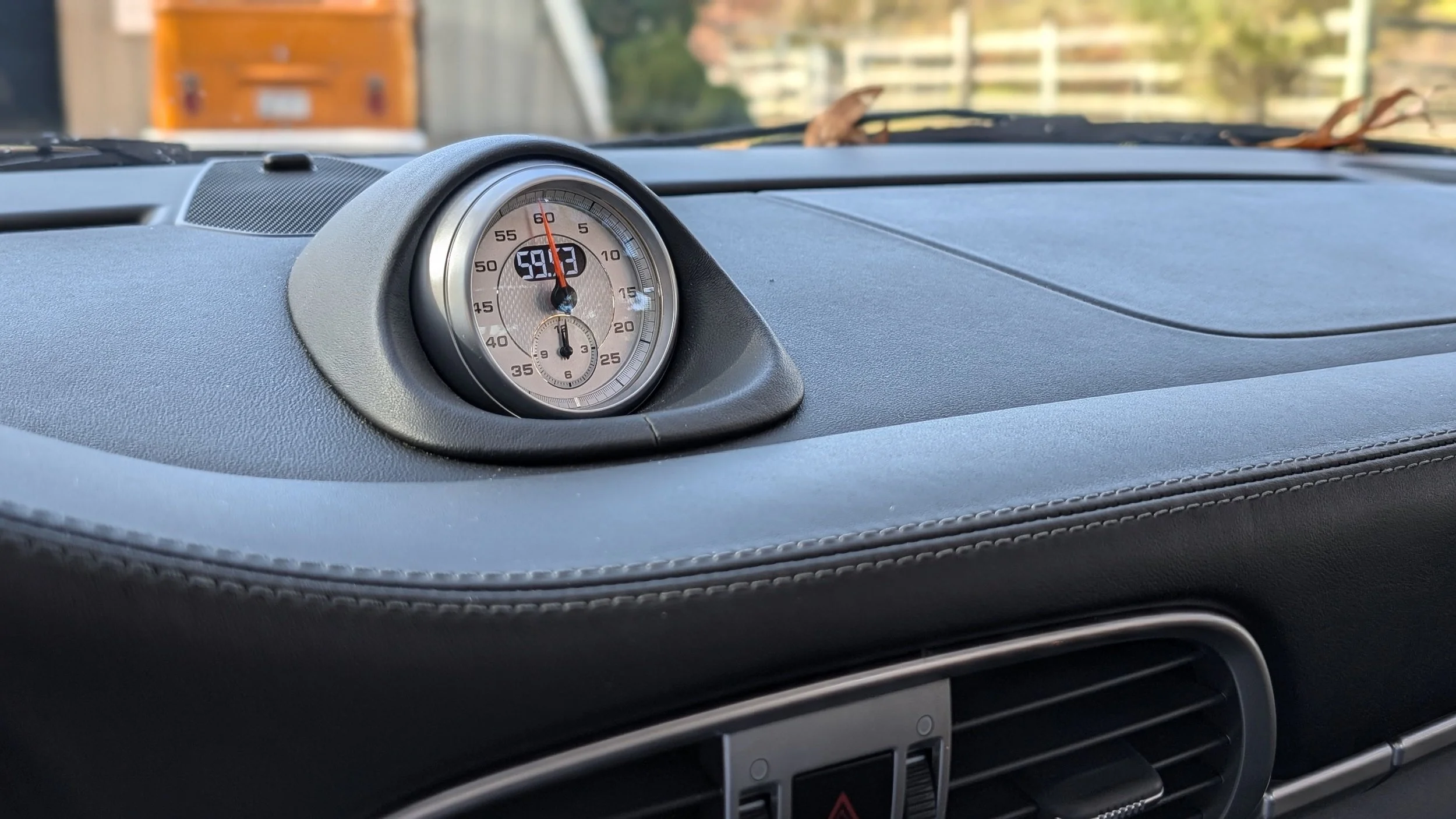 Close-up of a car's dashboard showing a central analog clock with a digital display, set against a blurred background of the windshield and trees outside.