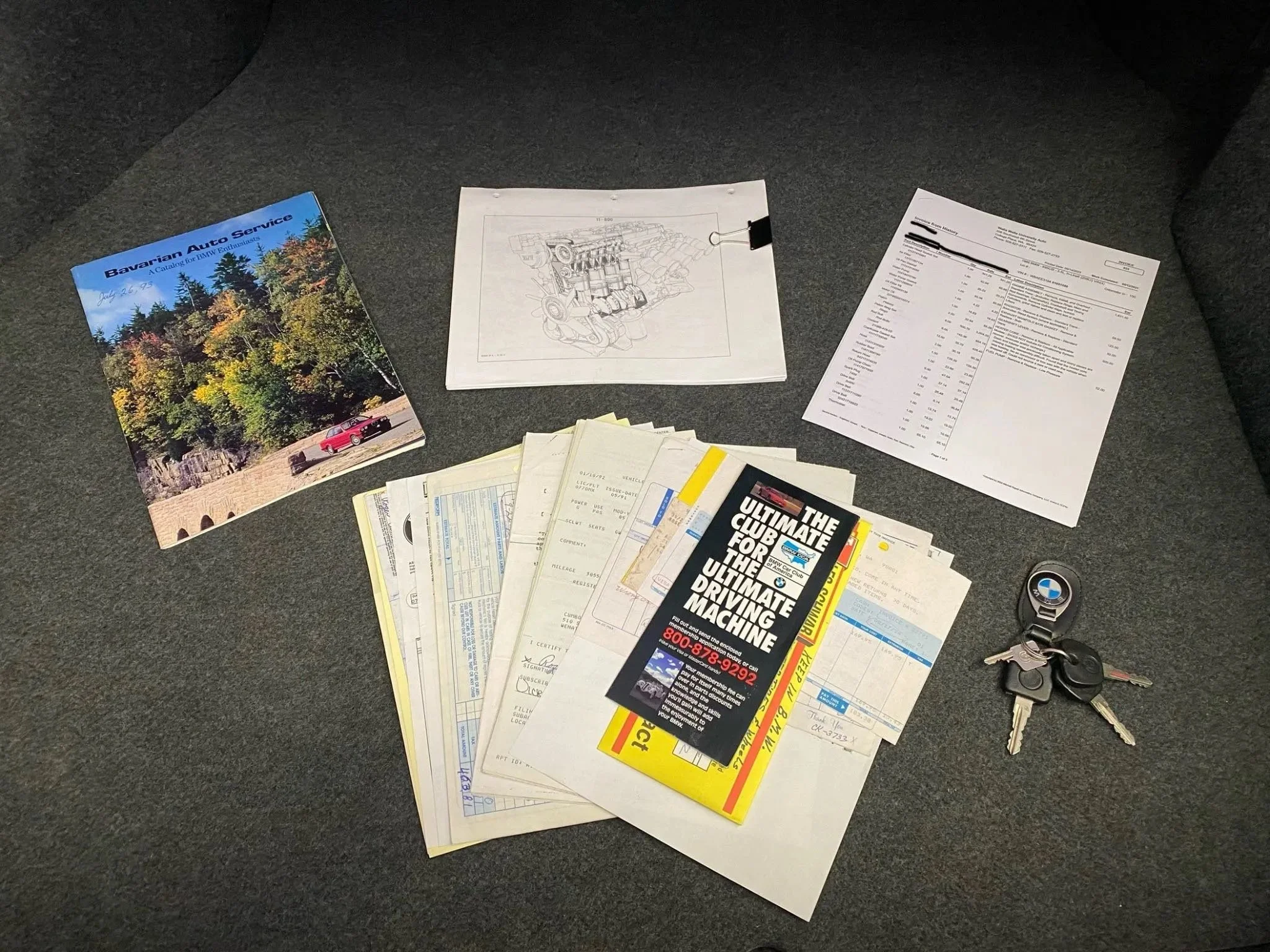 A collection of vehicle-related documents on a car's carpeted floor, including a BMW BMW brochure, a technical diagram, a detailed bill, a stack of registration papers, and a BMW keys with keychains.