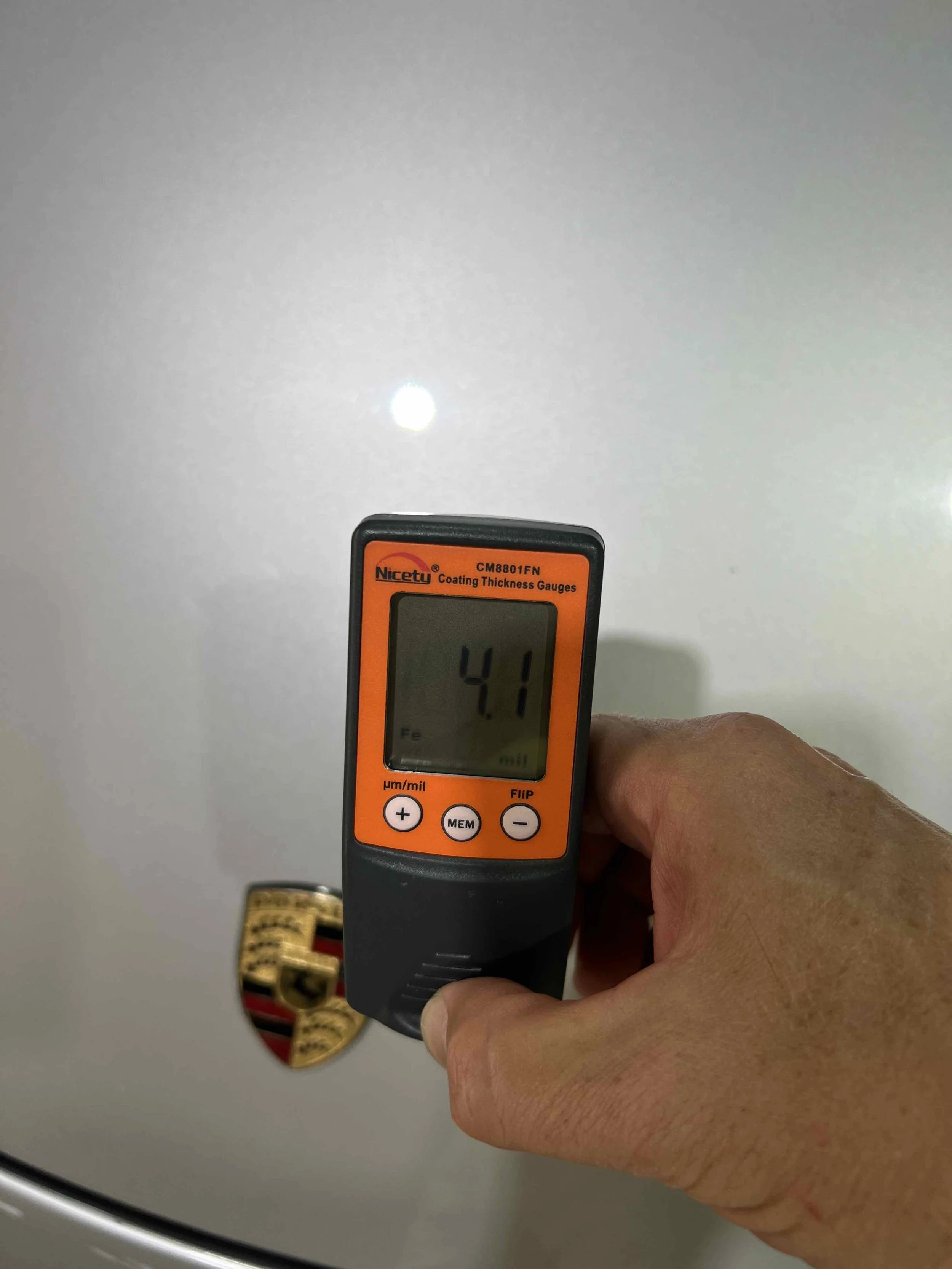 Hand holding a coating thickness gauge near a white surface with a logo.