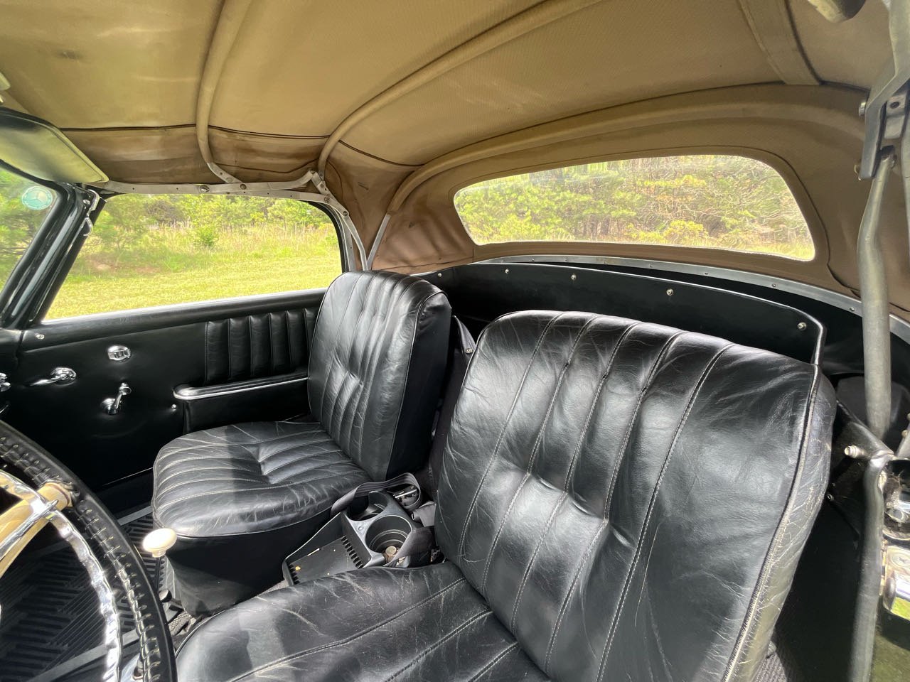 Interior of a vintage car with two black leather seats, a beige ceiling, and large windows showing greenery outside.