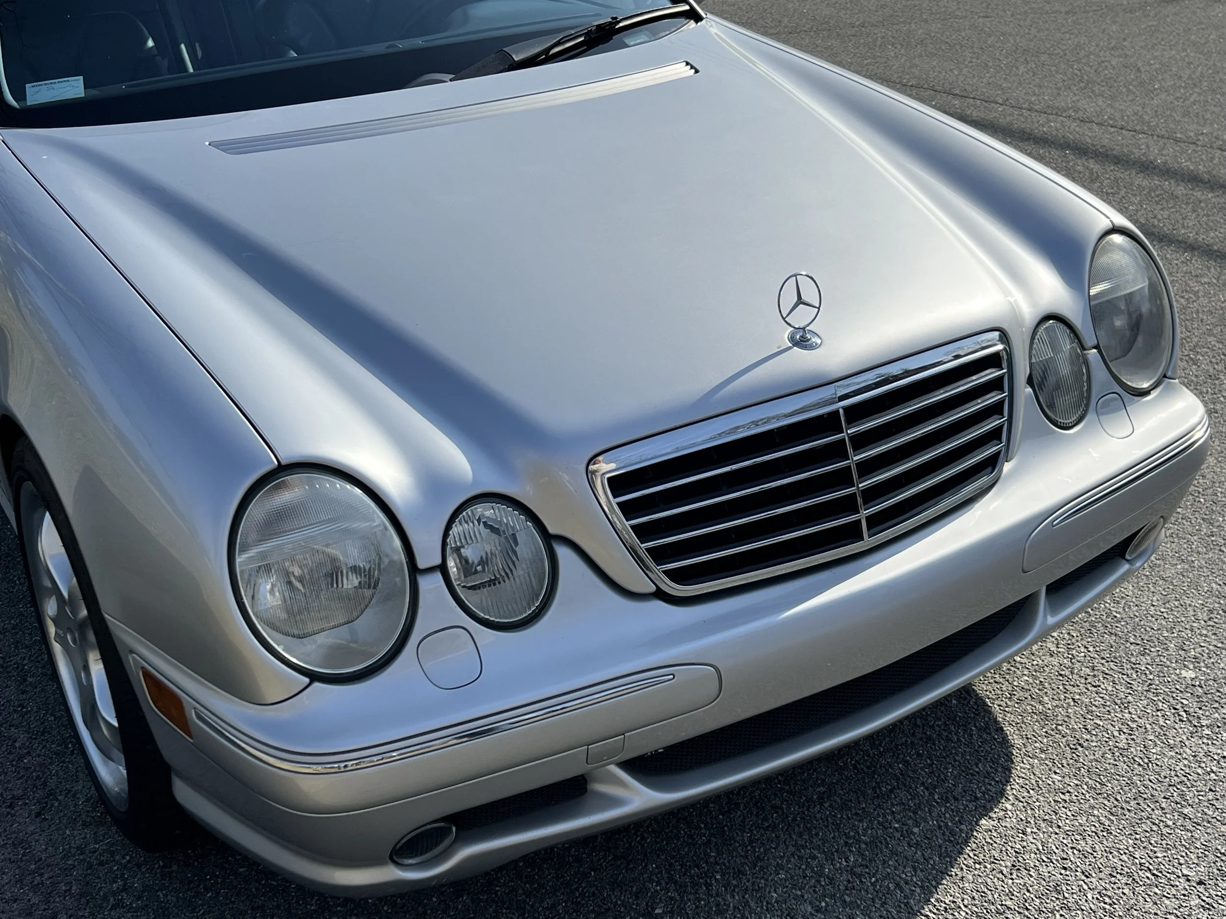A silver Mercedes-Benz car parked on a paved surface. The front of the car, including the grille, headlights, and hood ornament, is visible.