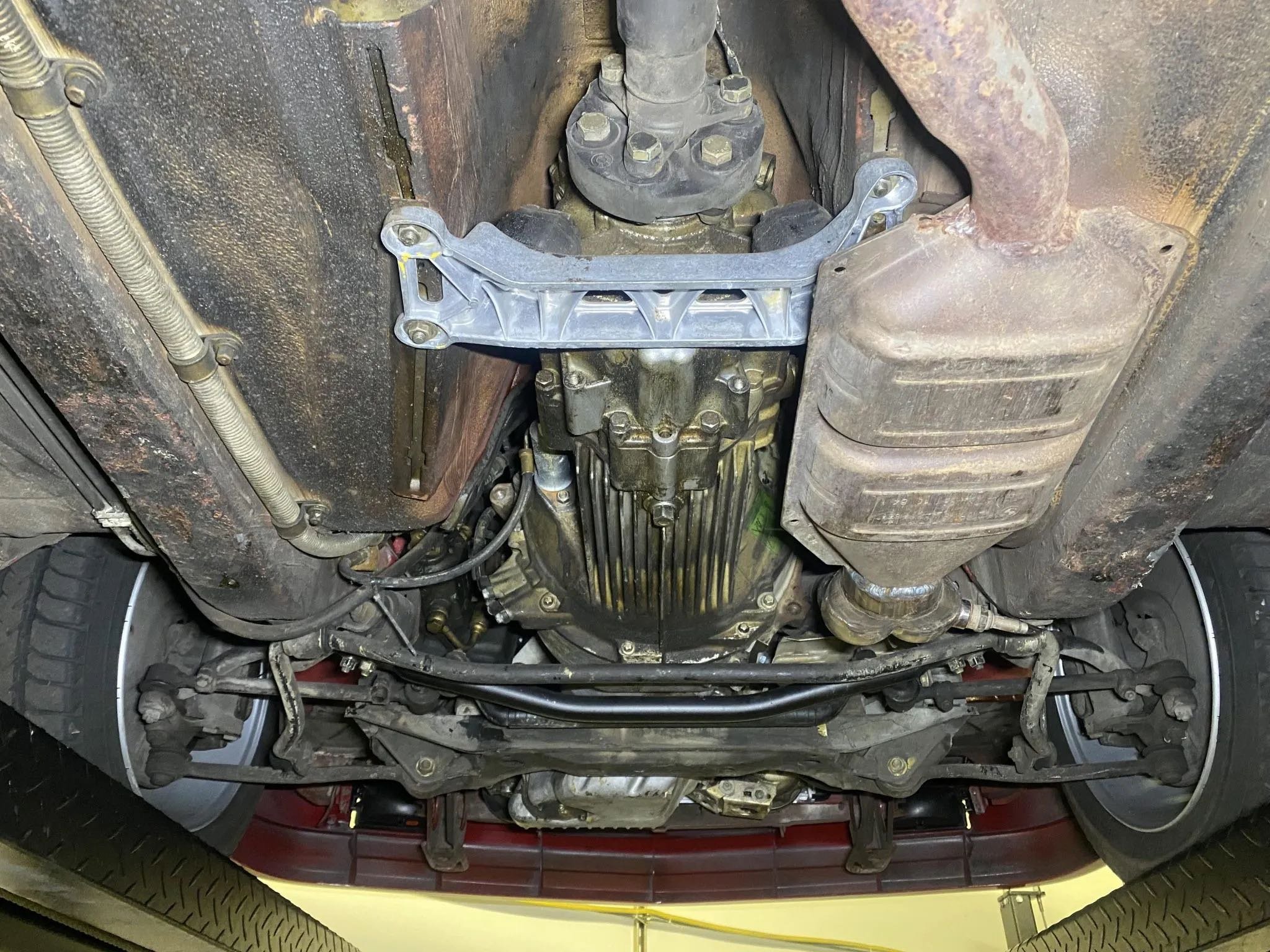 The image shows the underside of a car with a visible oil pan, suspension parts, and the engine valley, with a metal brace mounted to the engine components.