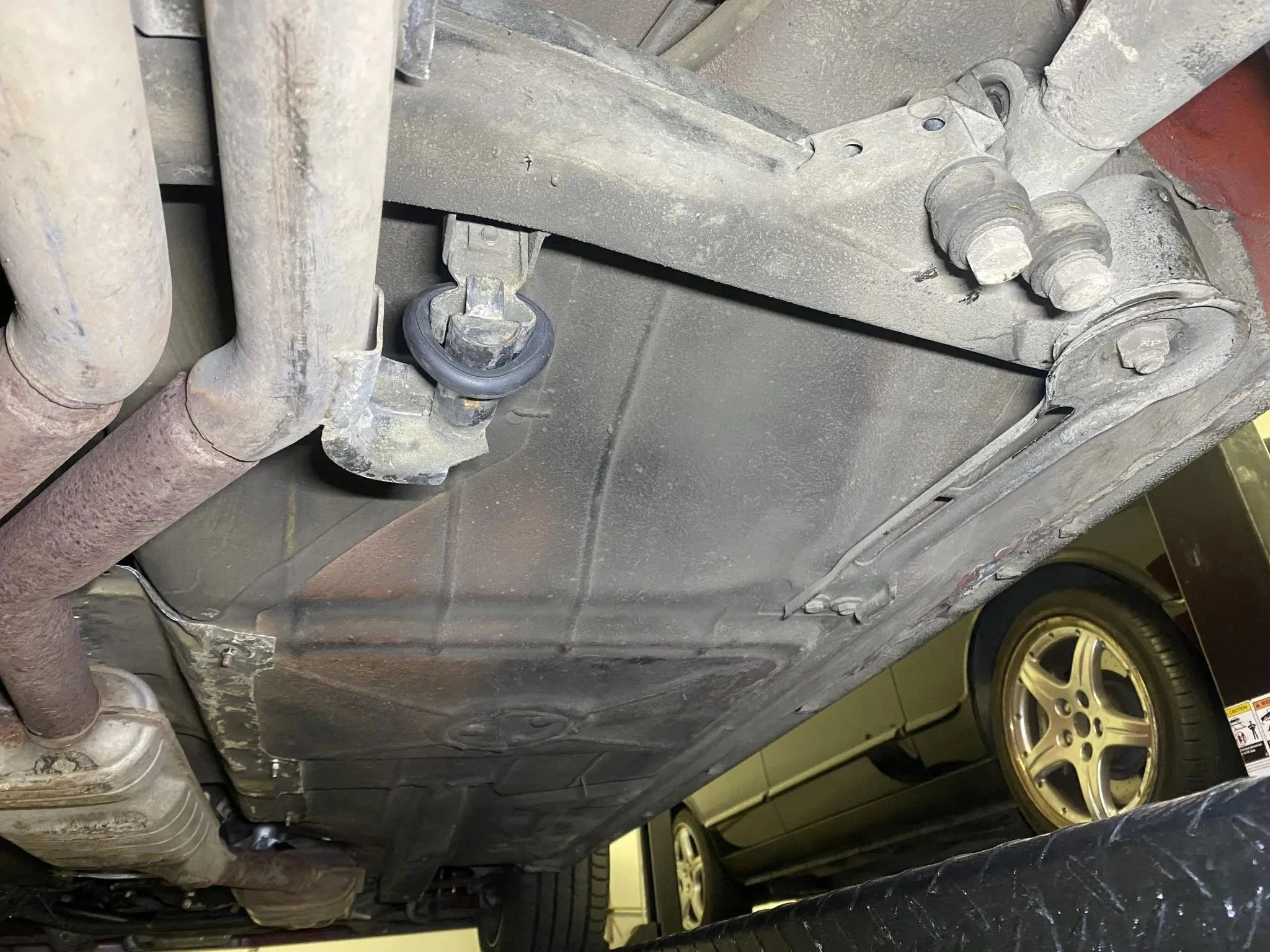 Underneath the car showing the metal frame, exhaust pipe, and various mechanical components, with a gold-colored wheel and tire visible in the background.
