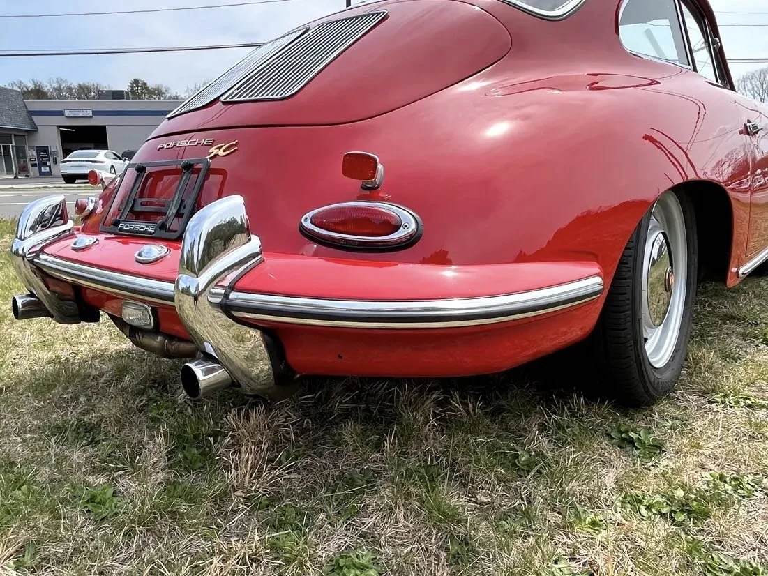 Rear view of a vintage red Porsche 911 Classic with chrome bumpers, a black license plate holder, and distinctive round taillights parked on grass.